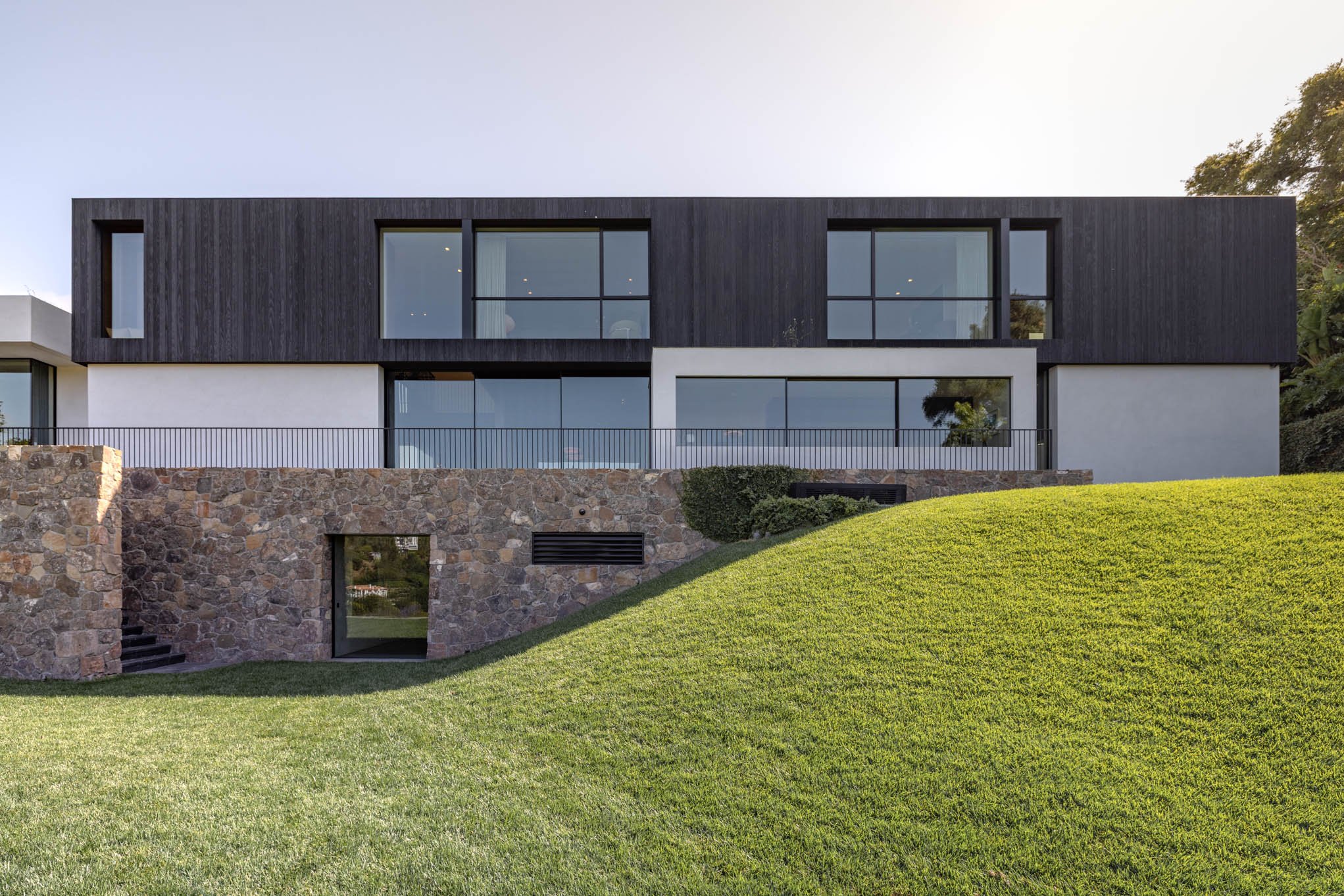 Modern house with black wooden upper section and white lower section, built on a sloped grassy yard with a stone foundation and a small underground door.
