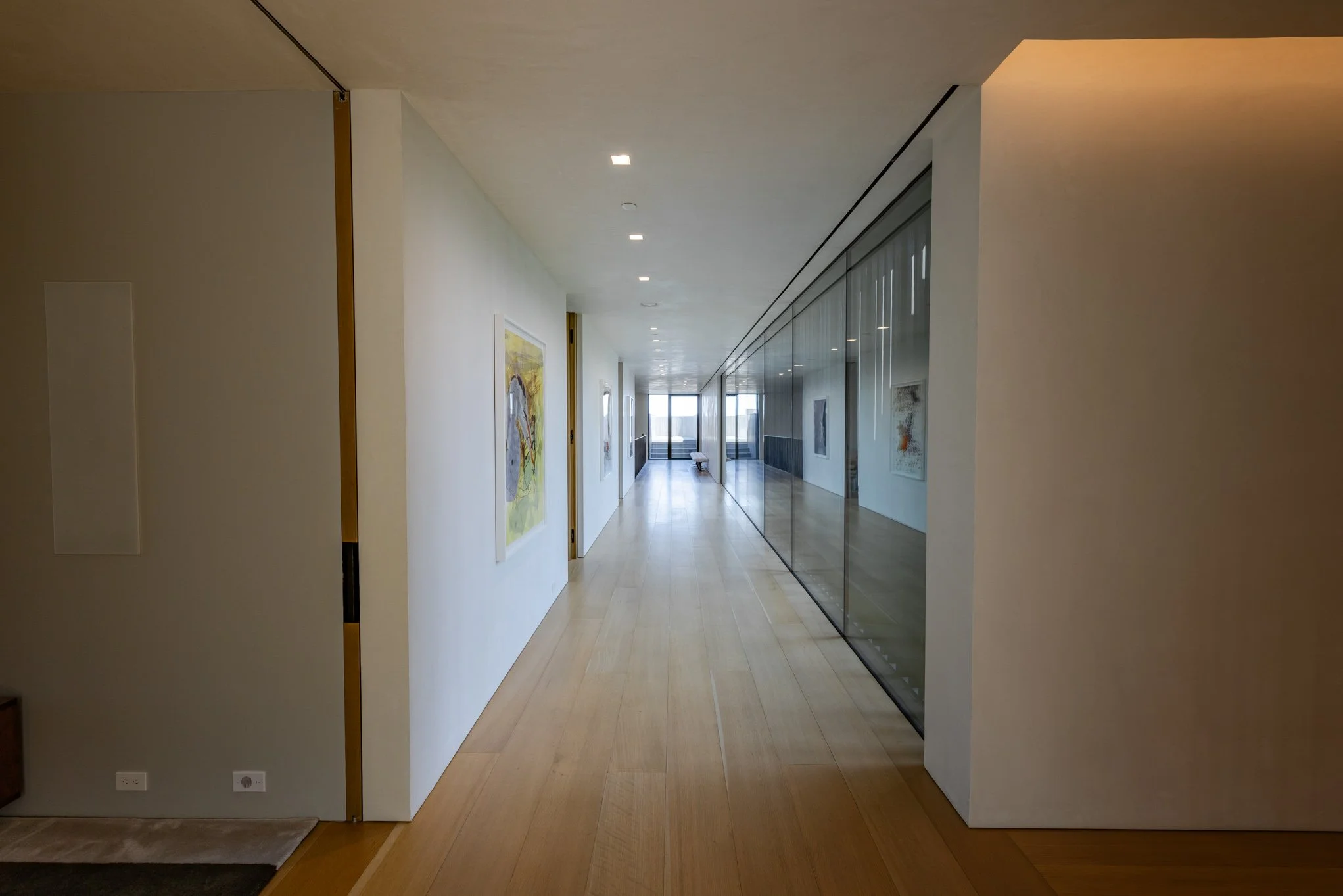 Modern hallway with wooden flooring, white walls, and a large glass partition on the right side, with artwork hanging on the left wall, leading to a bright room with large windows at the end.