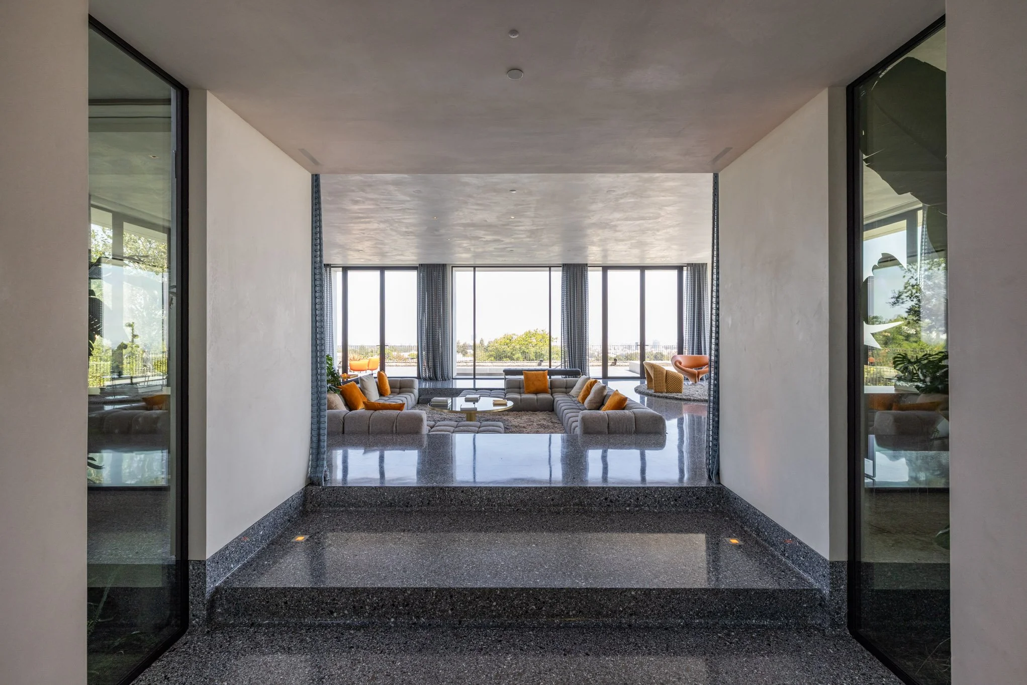 View from a modern entryway into a spacious living room with large windows, beige sectional sofa with orange pillows, and a view of greenery and blue sky outside.