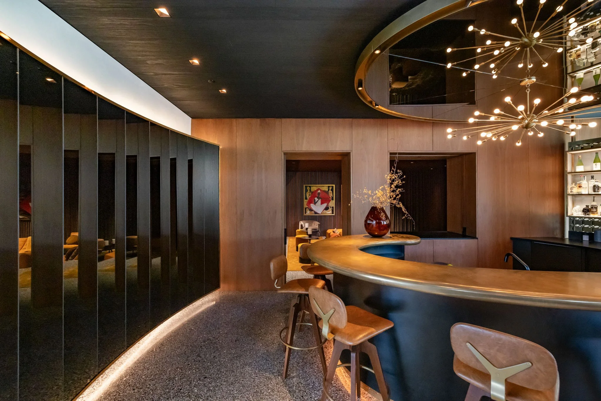 Modern hotel or restaurant bar with a curved black and wood bar counter, wooden bar stools, decorative lighting, and wooden wall paneling.