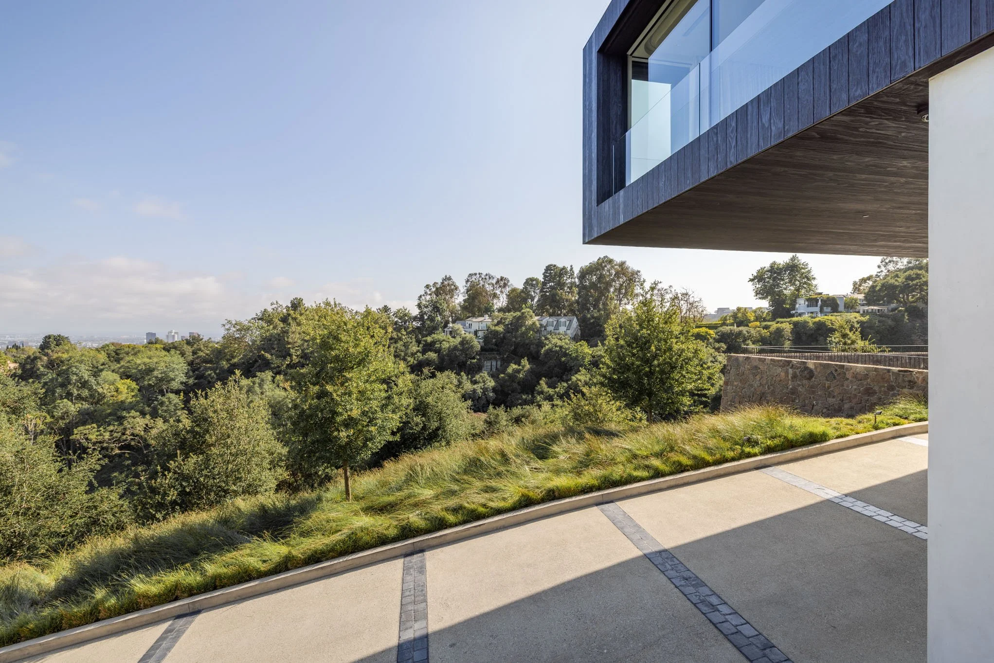 Modern house with a large overhanging balcony, overlooking a lush green landscape with trees and houses in the distance on a clear day.