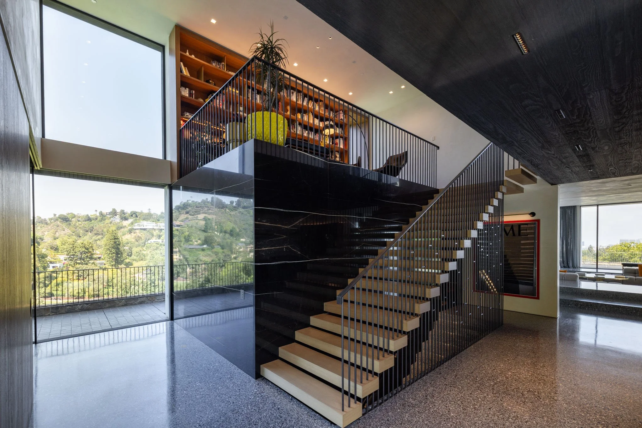 Modern interior staircase with black railings, open risers, and light wood treads, adjacent to large windows with a view of green hills, and a ceiling with dark wood panels.