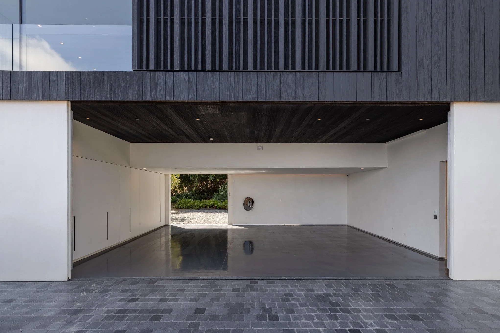Modern house garage with open door, black wooden upper facade, white interior, and greenery visible outside