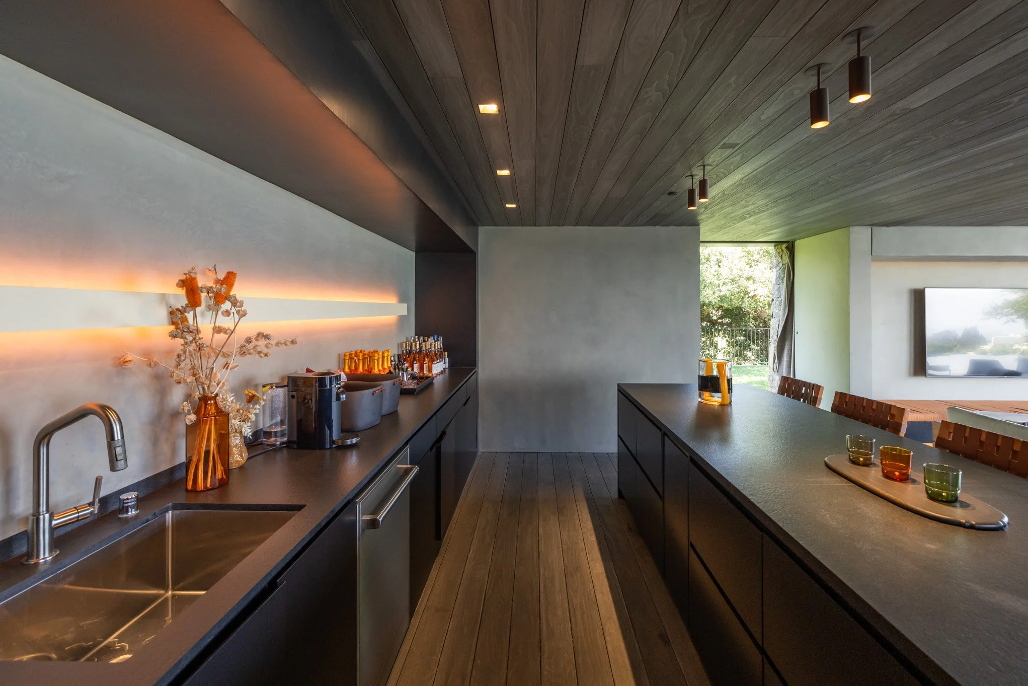Modern kitchen with black cabinets, wooden ceiling, and a large island with colorful glasses, and a view of a green outdoor space through a window.