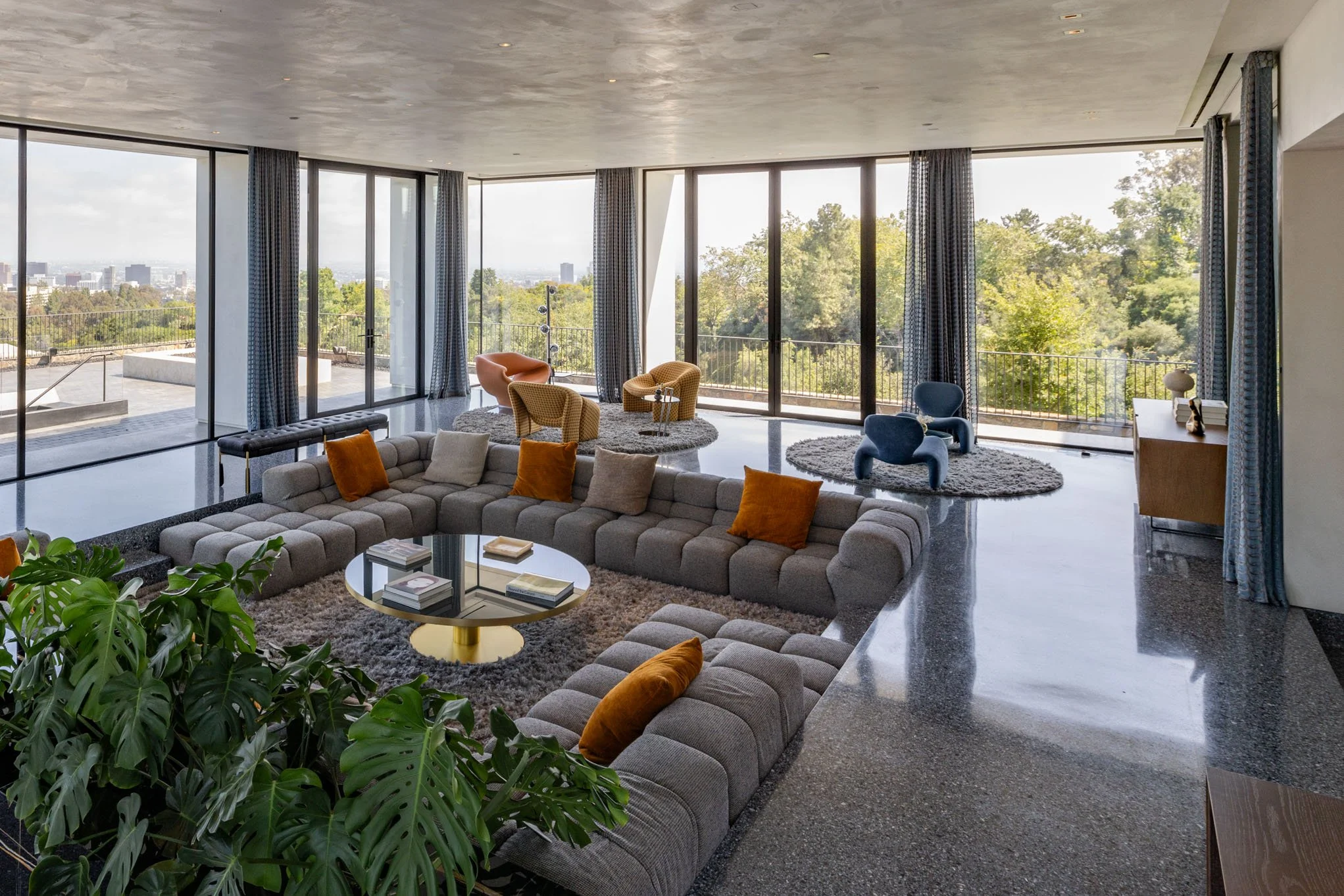 Modern living room with large floor-to-ceiling windows, a large gray sectional sofa with orange and gray pillows, a small black and gold circular coffee table with books, a textured plant in the corner, and various colorful armchairs on round rugs, w