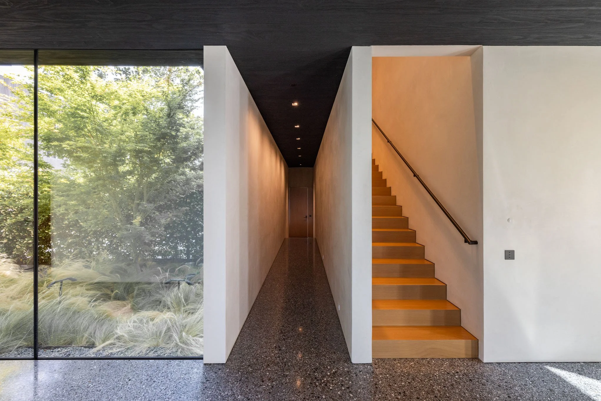 Modern interior hallway with a staircase on the right and a large window revealing greenery outside.
