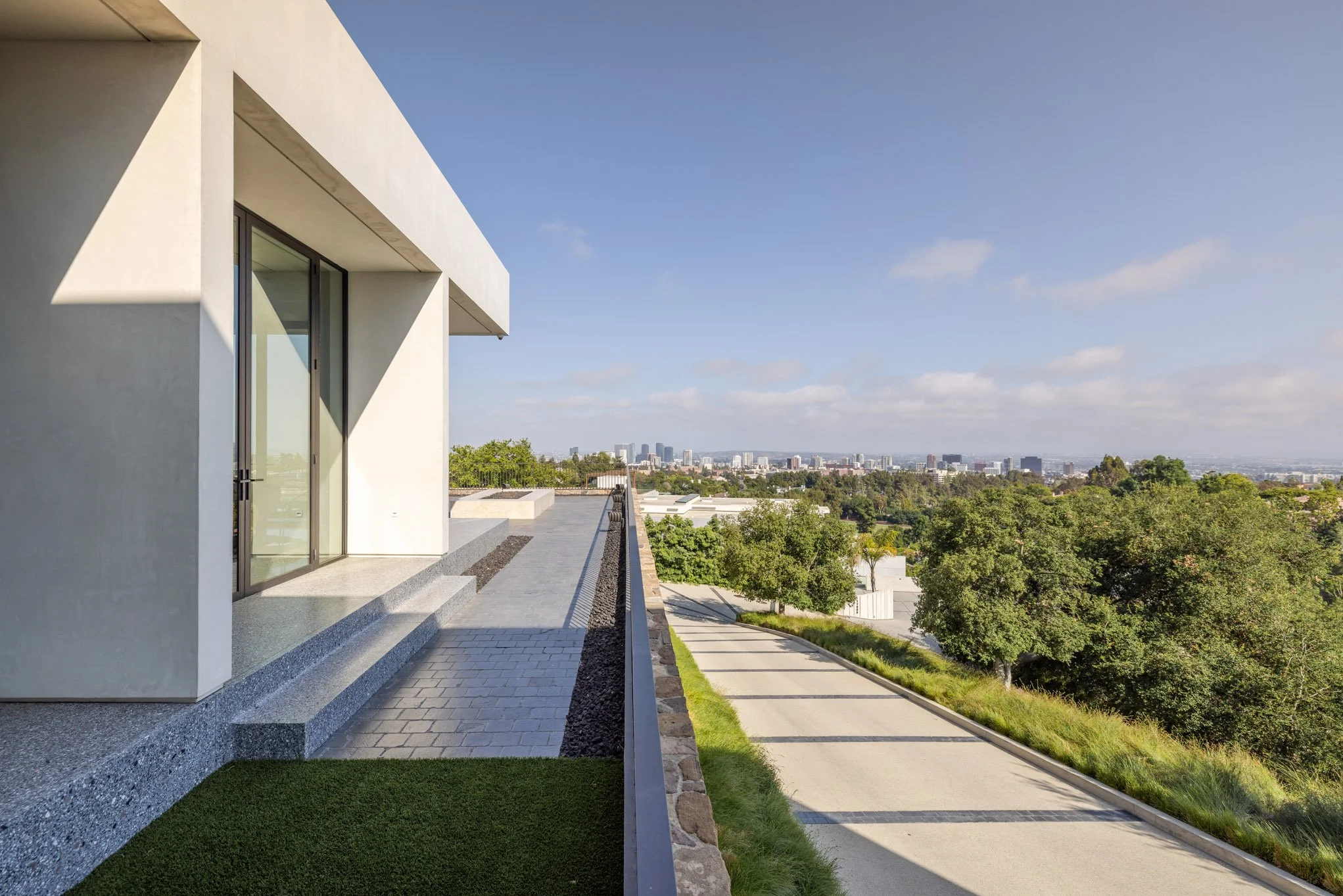 Modern house with a balcony overlooking a tree-lined street and cityscape in the distance under a partly cloudy sky.