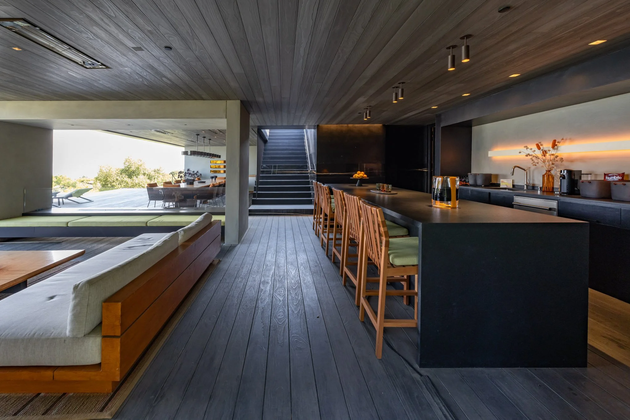 Modern open-concept kitchen and living area with wooden floors and ceiling, bar counter with chairs, and outdoor patio with dining area and lounge chairs
