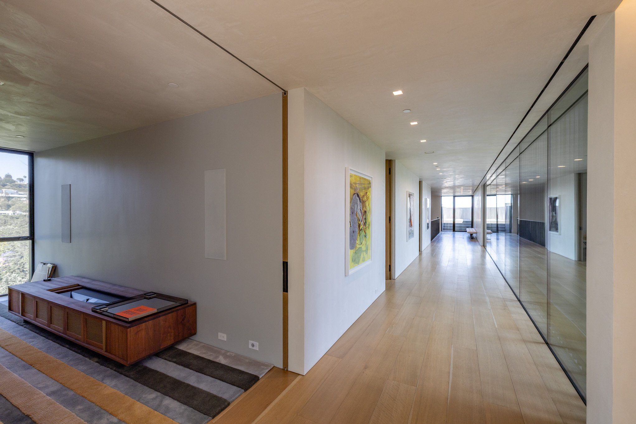 Modern hallway with wooden flooring, white walls decorated with colorful artwork, and a large glass partition on the right reflecting the hallway. At the end, there is a sitting area with large windows letting in natural light.