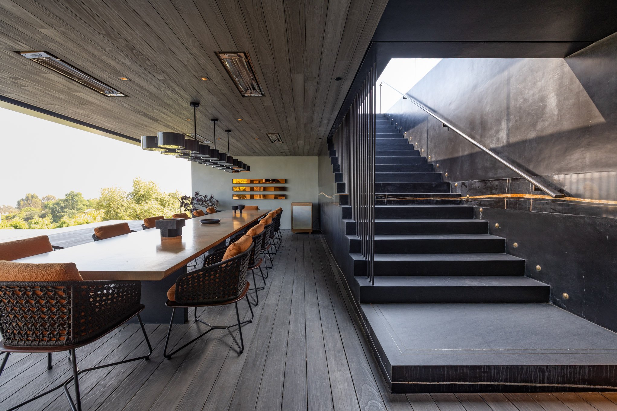 Modern room with a long wooden dining table, black chairs with beige cushions, a staircase with black steps and a metal railing, wooden and black ceiling, and large window with a view of green trees outside.