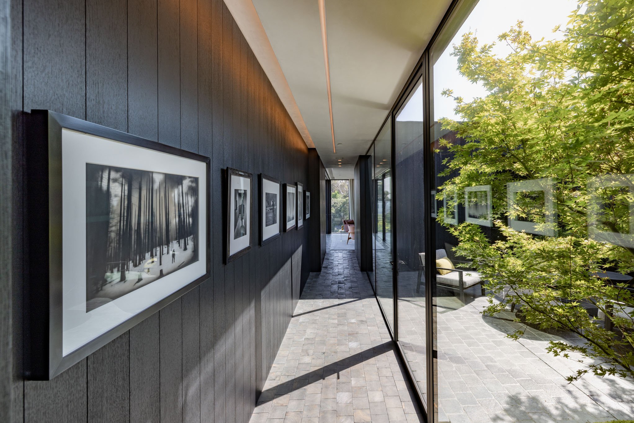 Indoor hallway with black wall decorated with framed black and white photographs on the left and large glass windows on the right, revealing a patio with outdoor furniture and green trees outside.