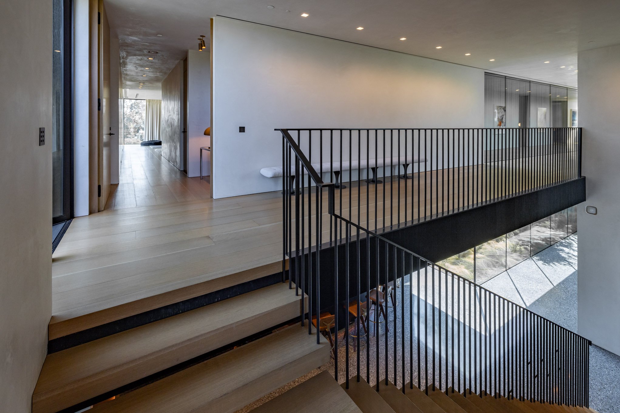 Modern interior home with wooden stairs, black railing, white walls, ceiling lights, and a view of a sitting area with large windows.