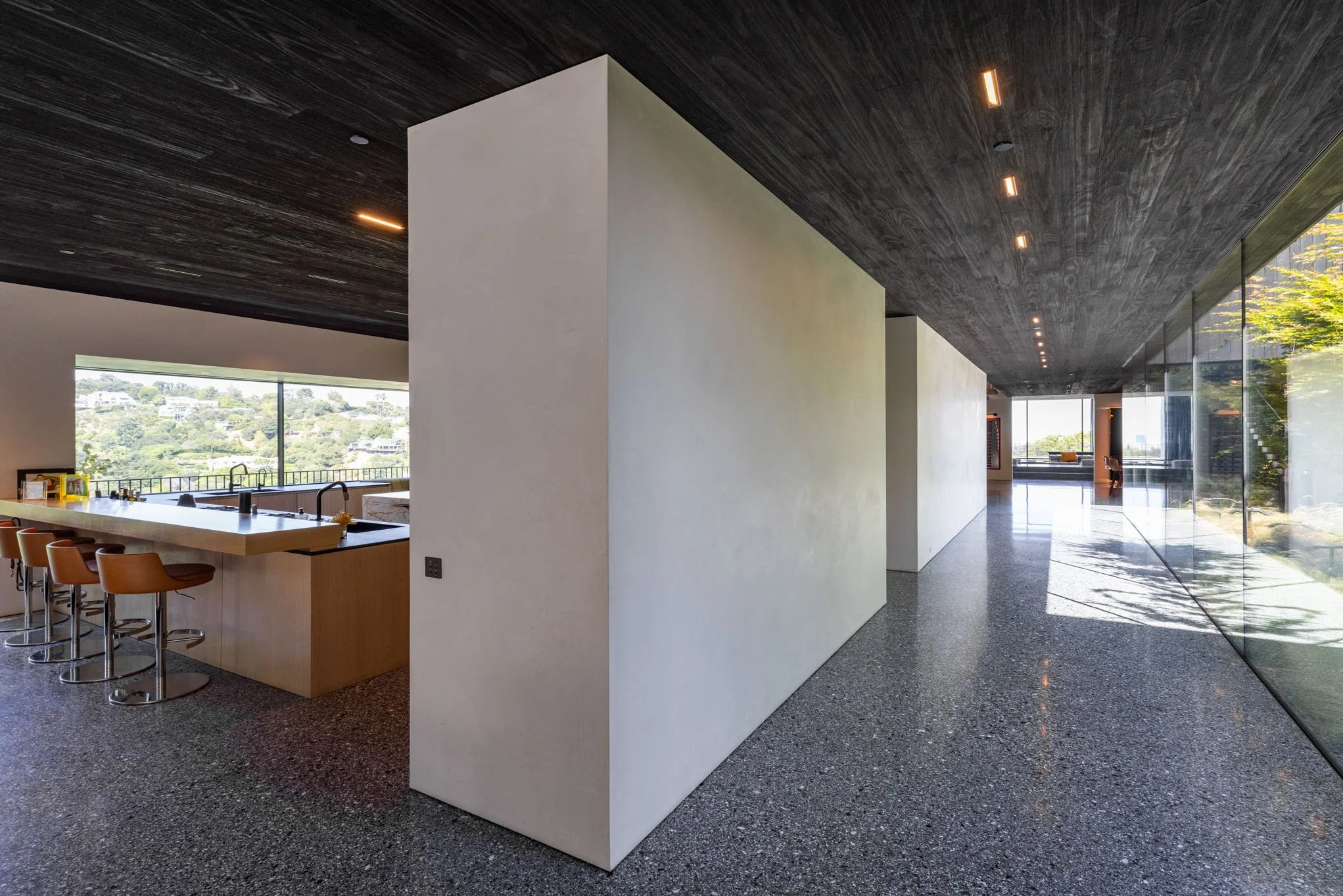 Modern interior of a house with a kitchen island, barstools, large windows showing scenic view, white walls, black textured ceiling, and glass walls with outdoor greenery.