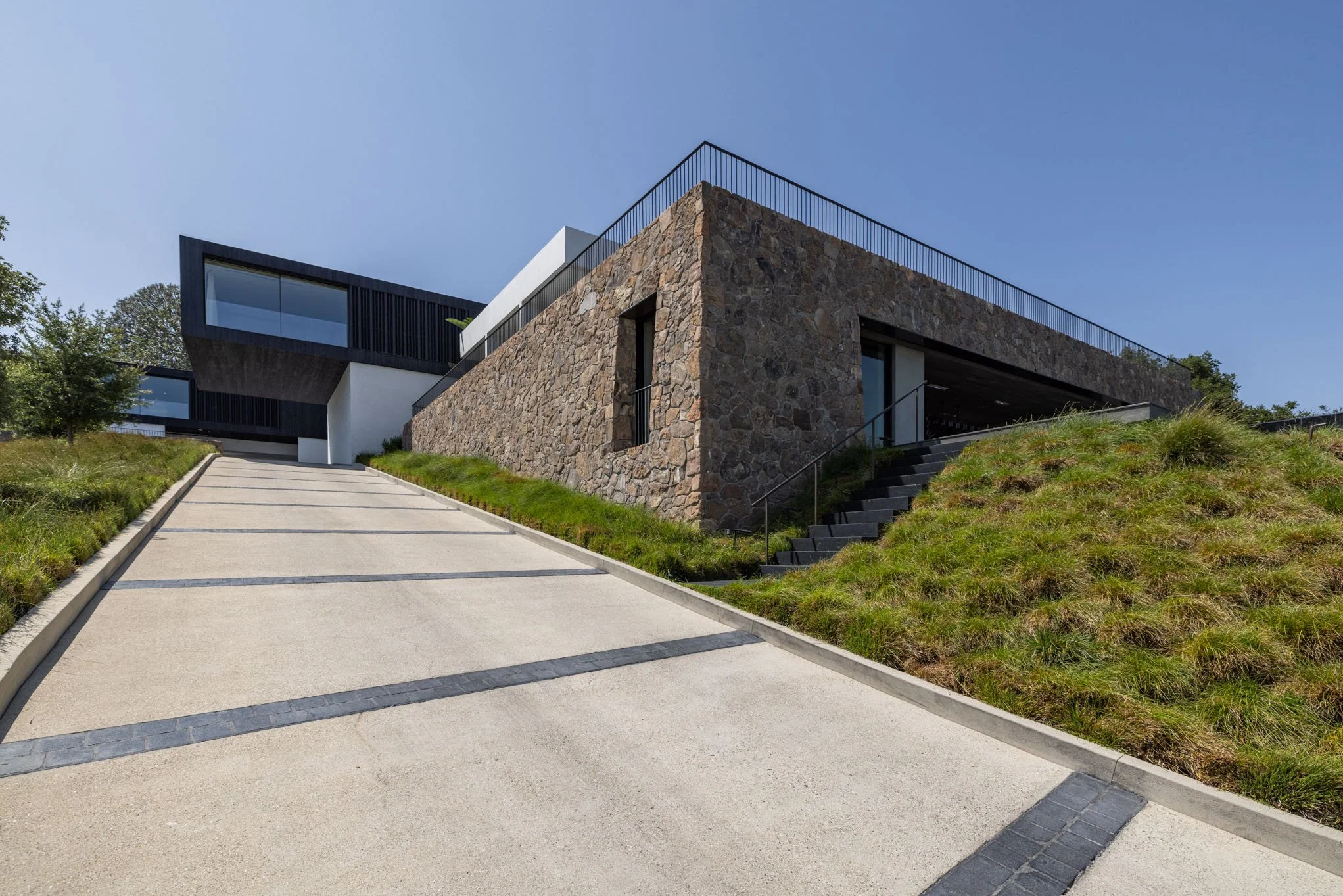 Modern house with a stone and white exterior, set on a hillside with a inclined driveway and stairs, greenery, and a clear blue sky.