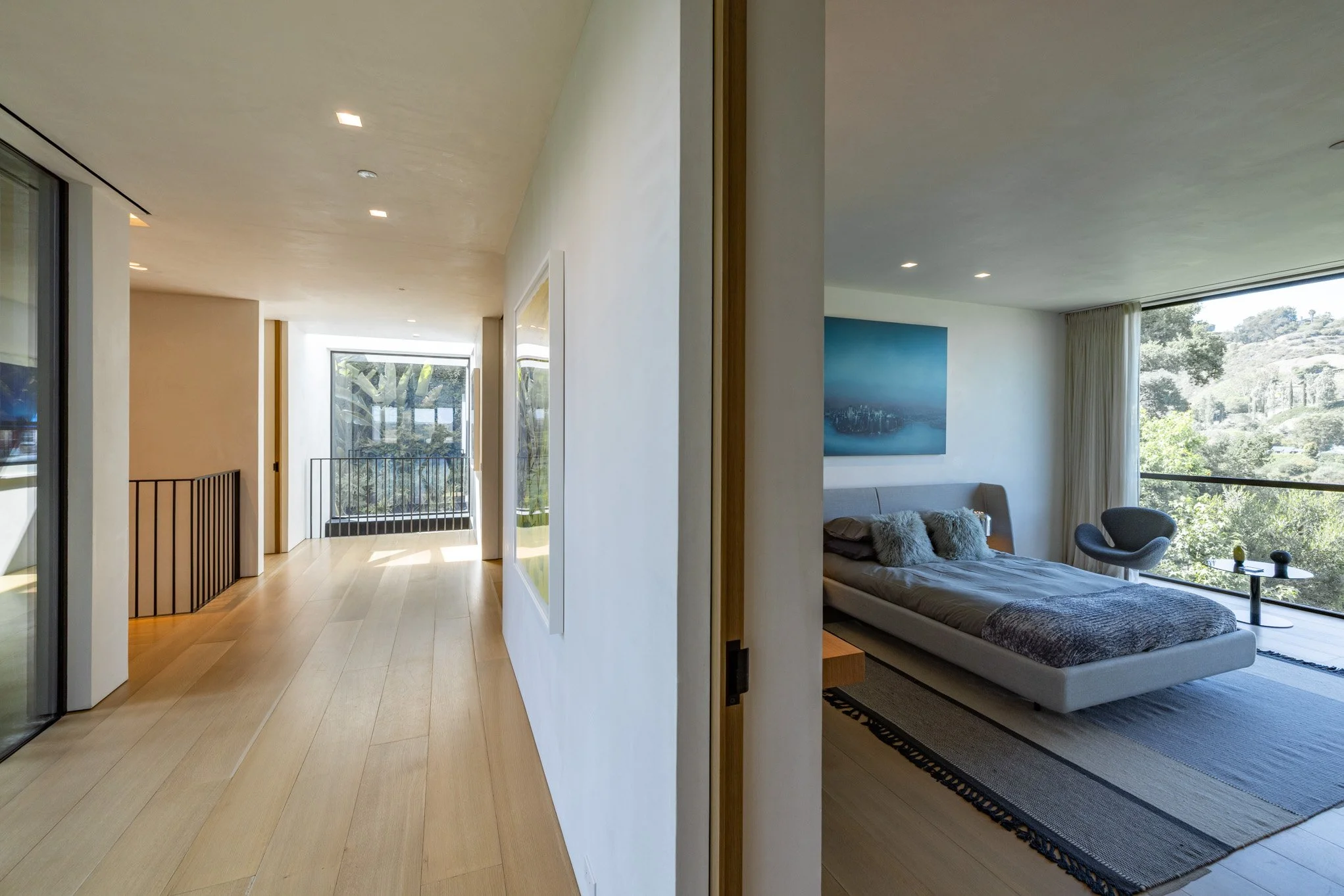 Split view of a modern house interior showing a hallway with wooden flooring and a bedroom with large window views of trees and landscape.