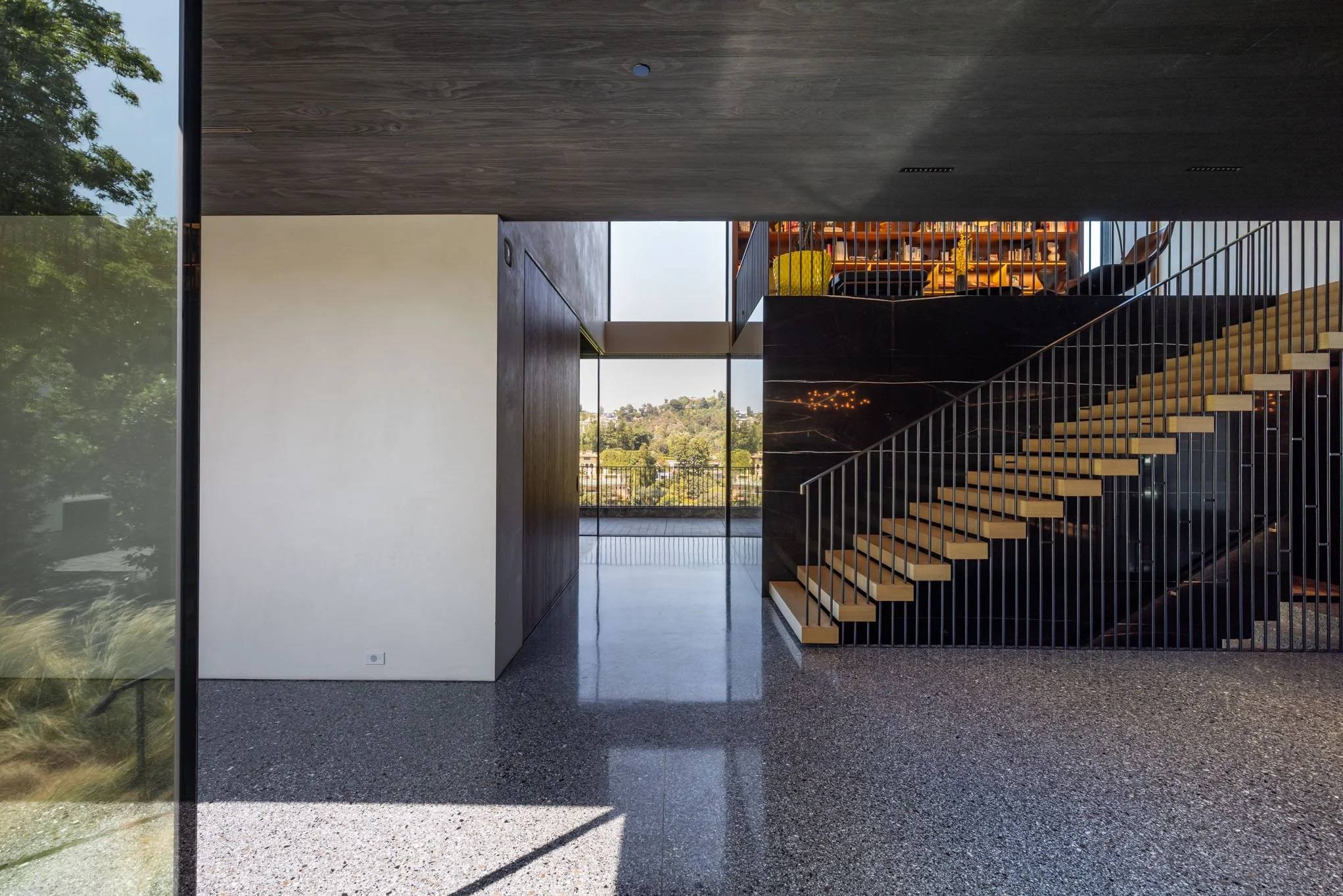 Modern interior with a staircase featuring light wood steps and black metal railing, leading to an upper level with visible shelving and seating. Glass doors reveal an outdoor view of trees and a cityscape.