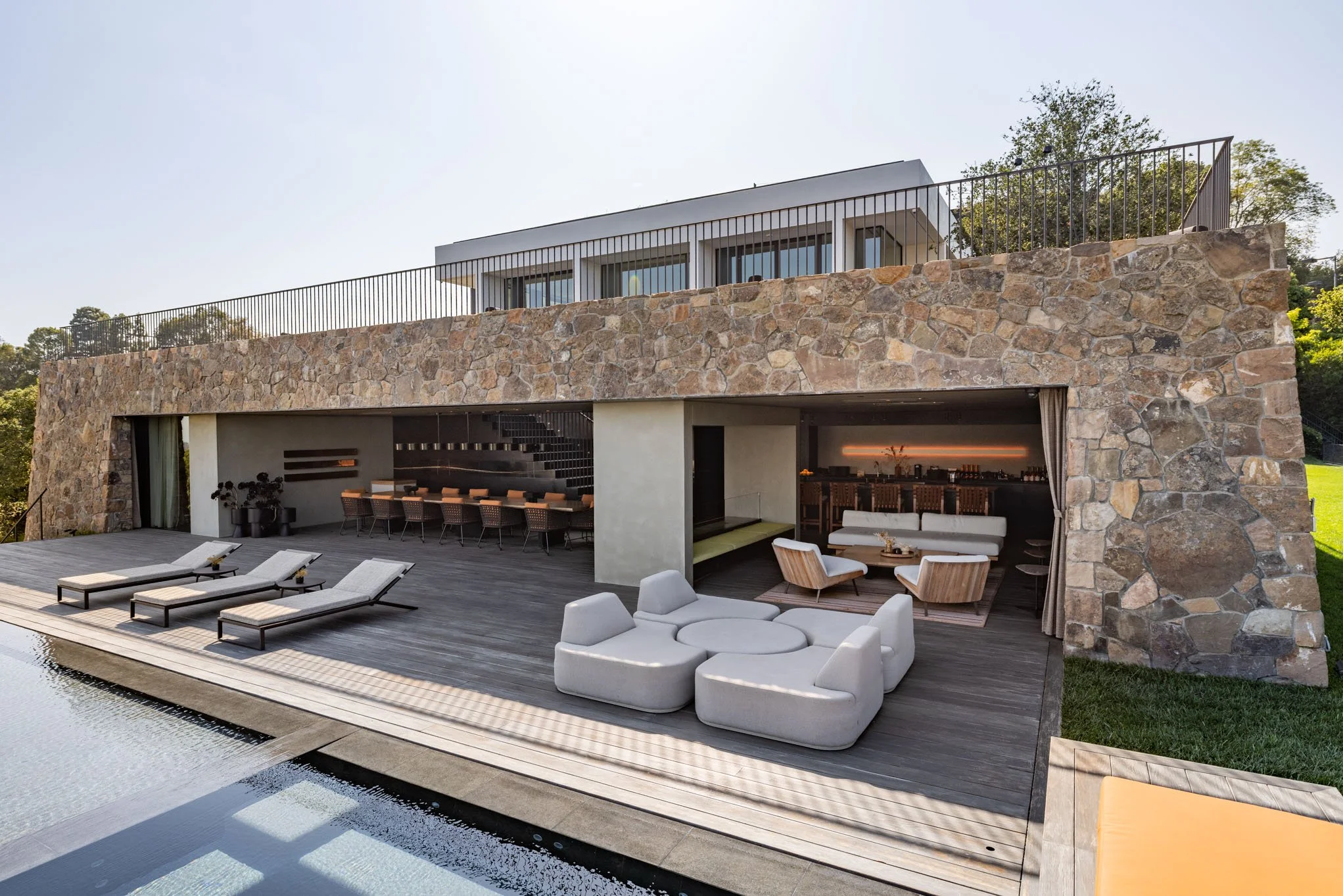 Modern house with stone facade and outdoor pool area with lounge chairs, cushioned seating, and a covered lounge space.