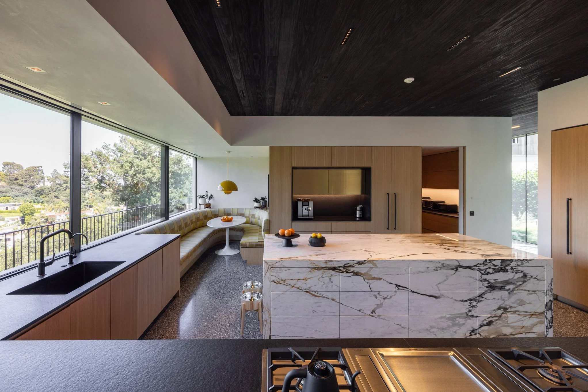 Modern kitchen with large windows, a marble island, and a cozy circular seating area with striped cushions and a yellow hanging light.