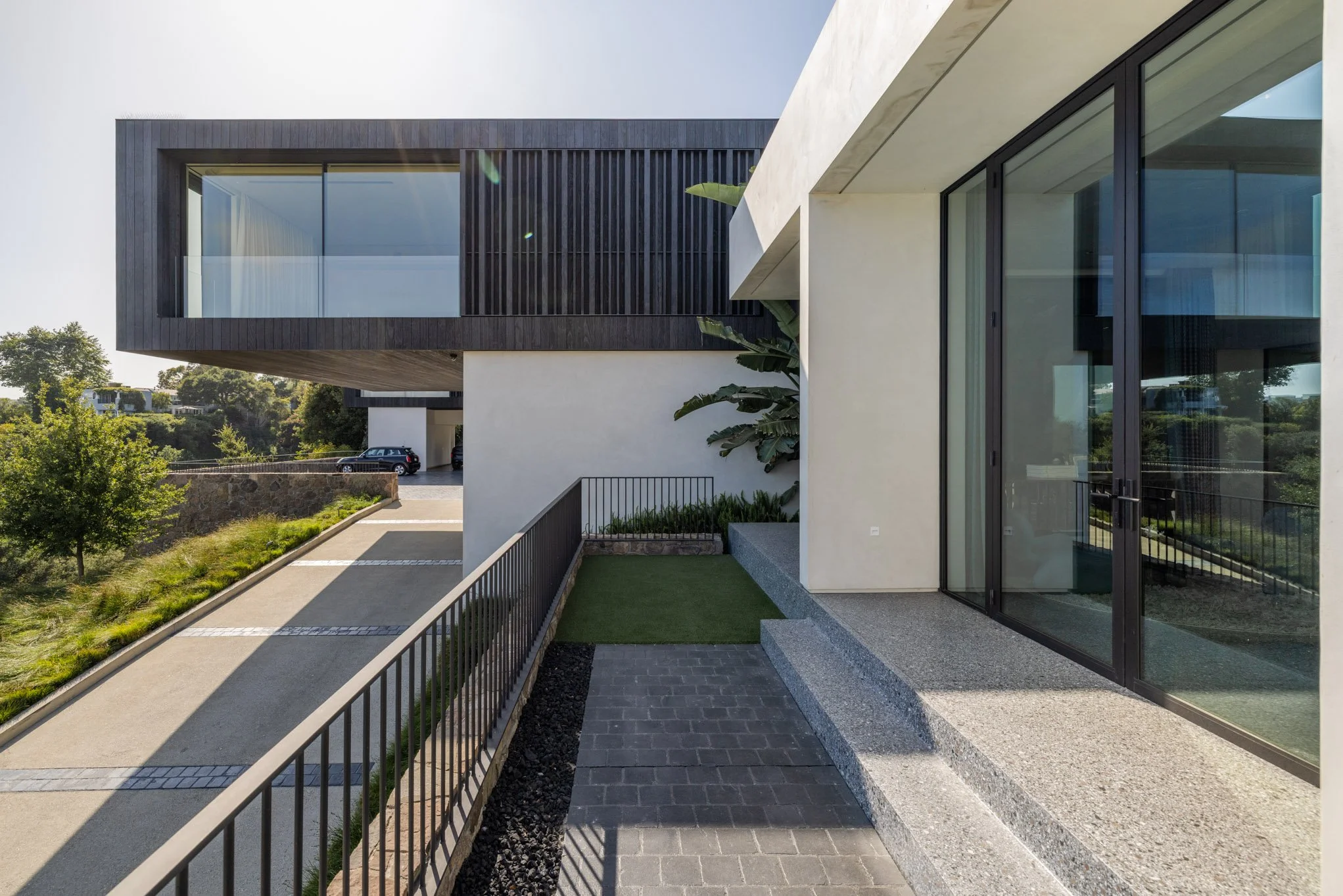 Modern residential building with large glass sliding doors and a balcony with black wooden paneling, situated in a landscaped area with trees and a driveway.