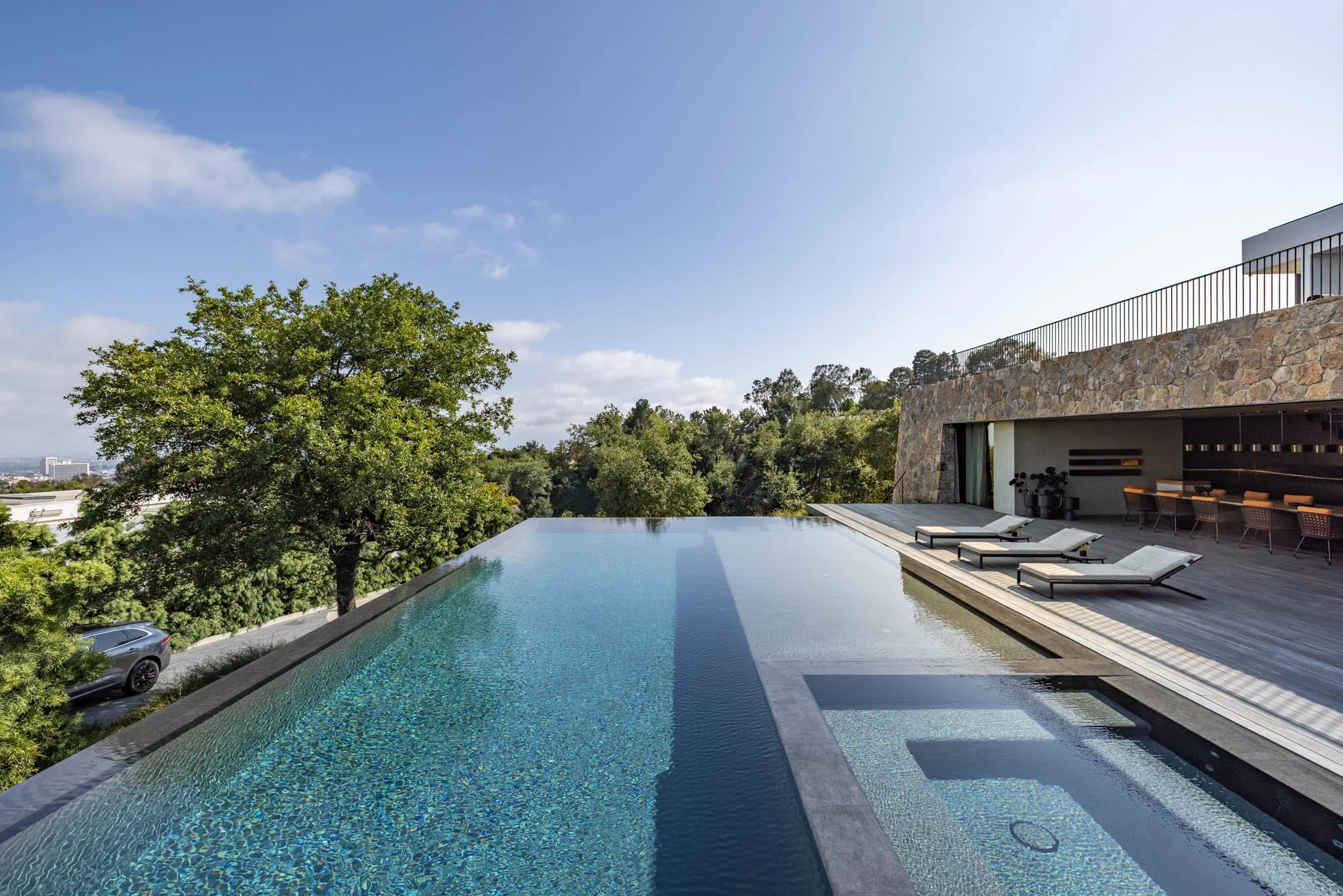 Luxury modern infinity pool overlooking trees and city skyline with lounge chairs on wooden deck and outdoor seating area at a contemporary house.
