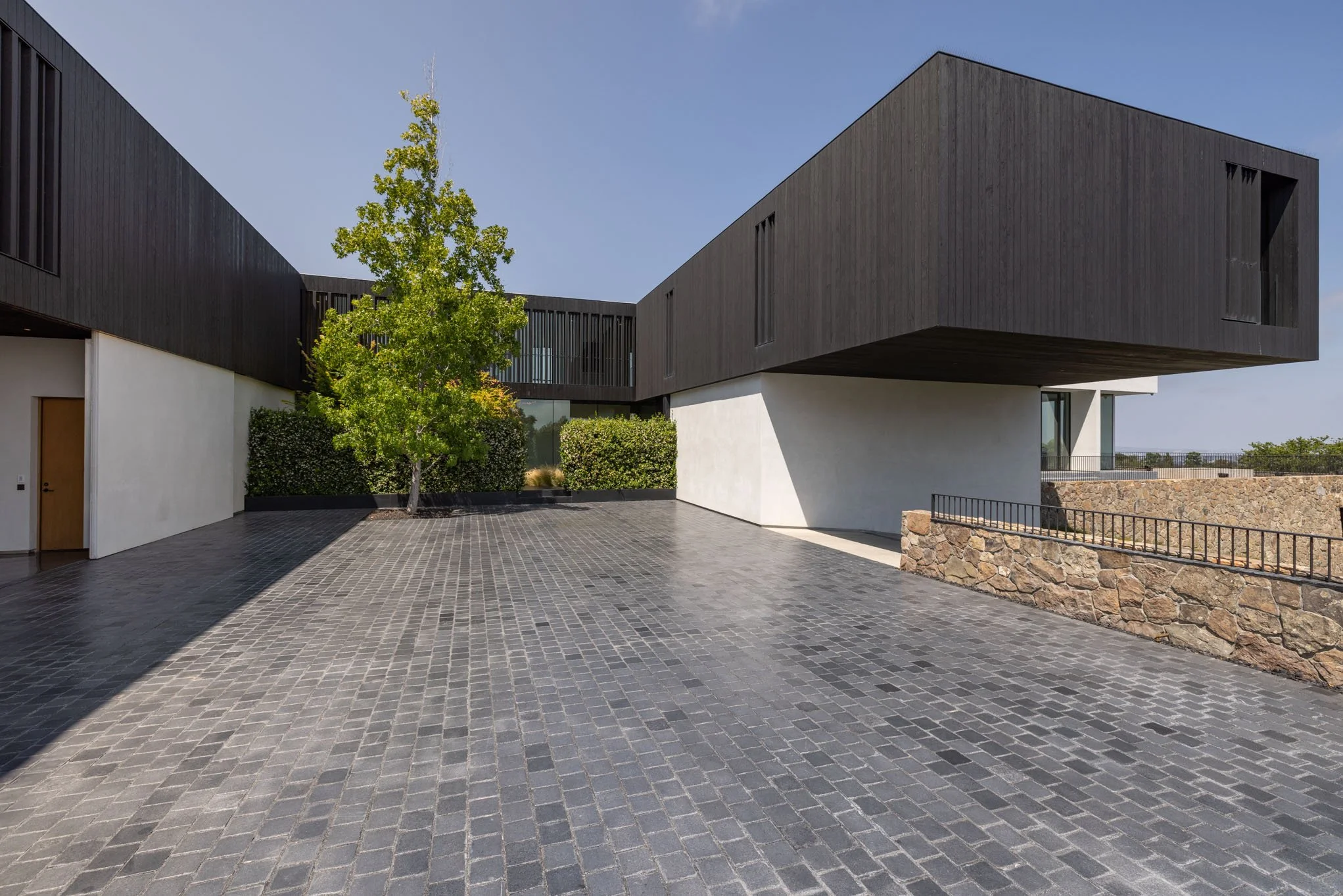 Modern building with black wooden exterior, white concrete section, stone wall, paved courtyard, and a green tree under a clear blue sky.