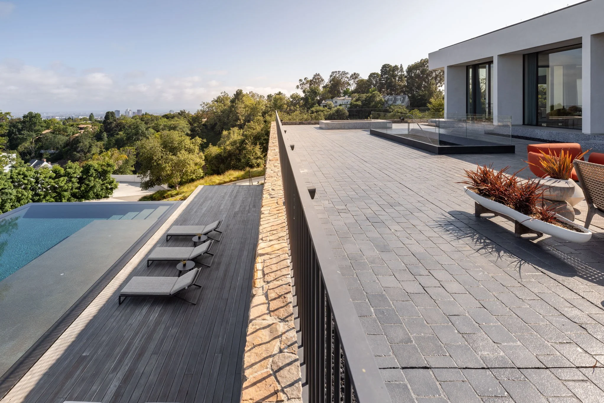 Modern rooftop terrace with outdoor seating, potted plants, and a pool, overlooking a green landscape with trees and distant buildings.
