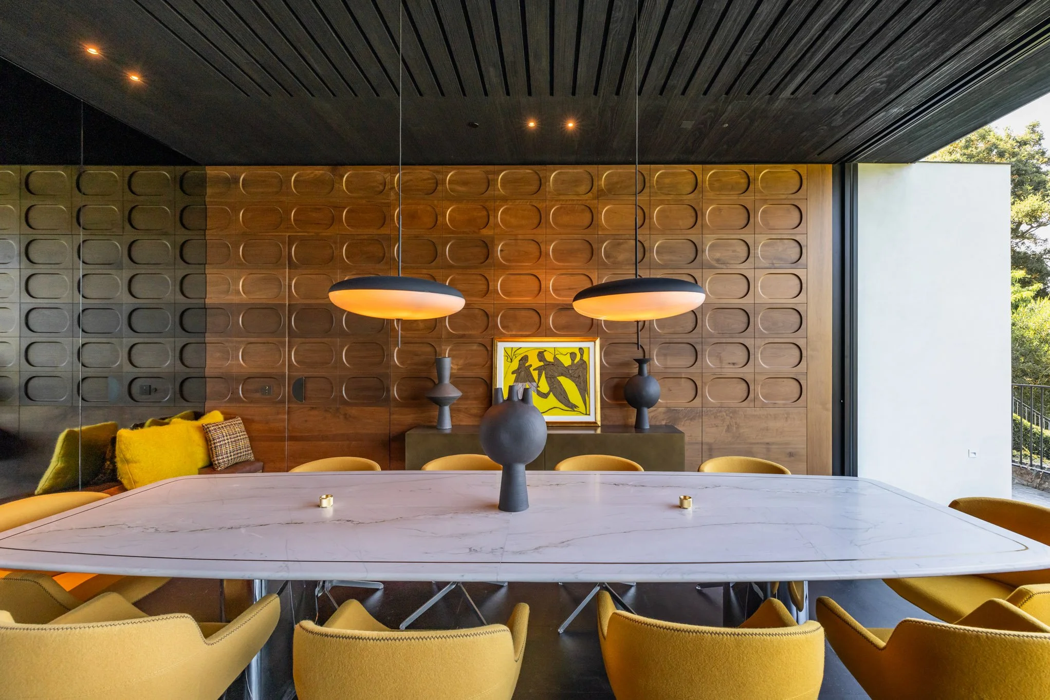 Modern dining room with a white marble table surrounded by yellow chairs, a wooden accent wall, black pendant lights, and yellow art on the wall.
