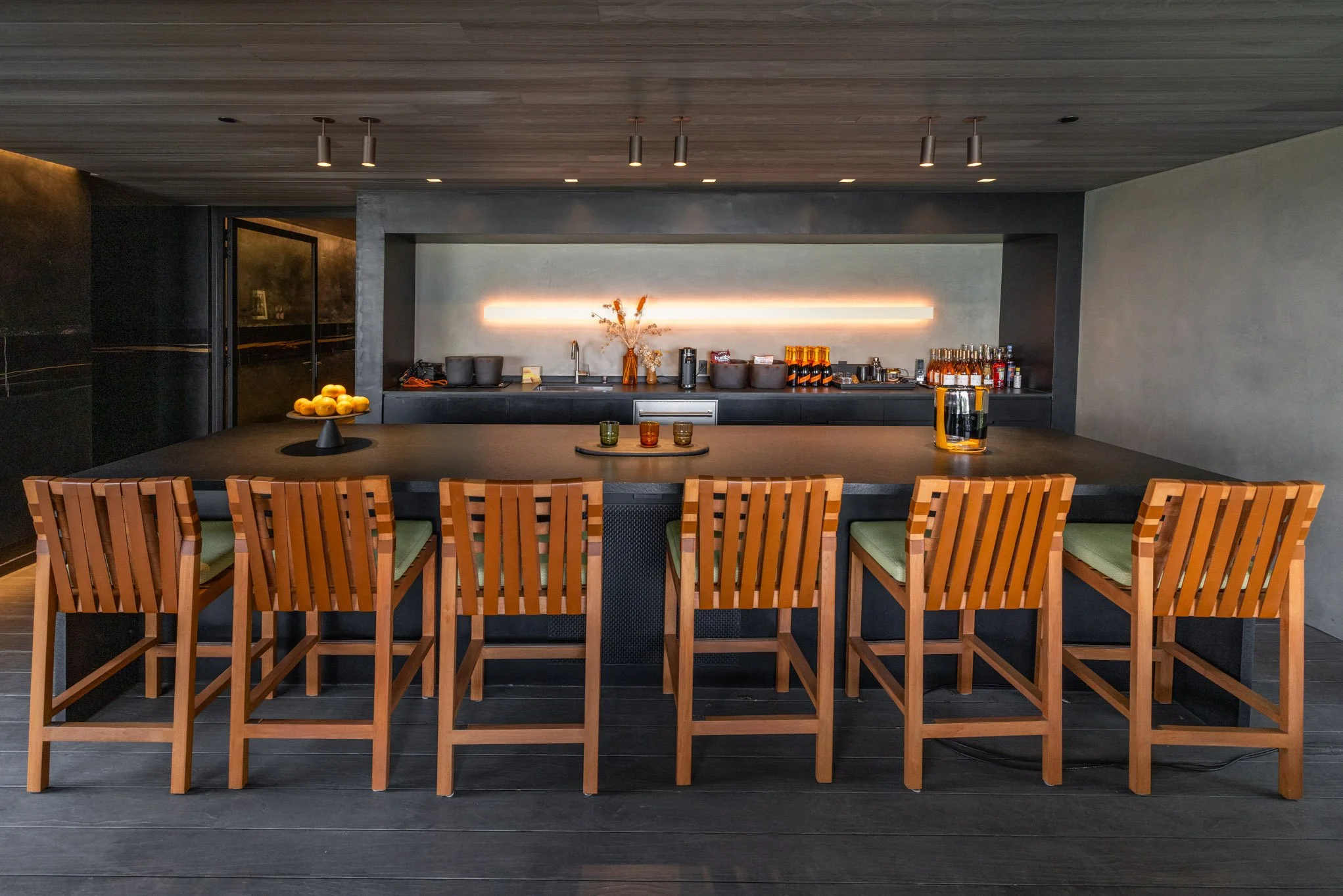 Modern bar with a dark countertop, wooden barstools with green cushions, and a minimalist backlit wall behind the bar area stocked with drinks and glassware.