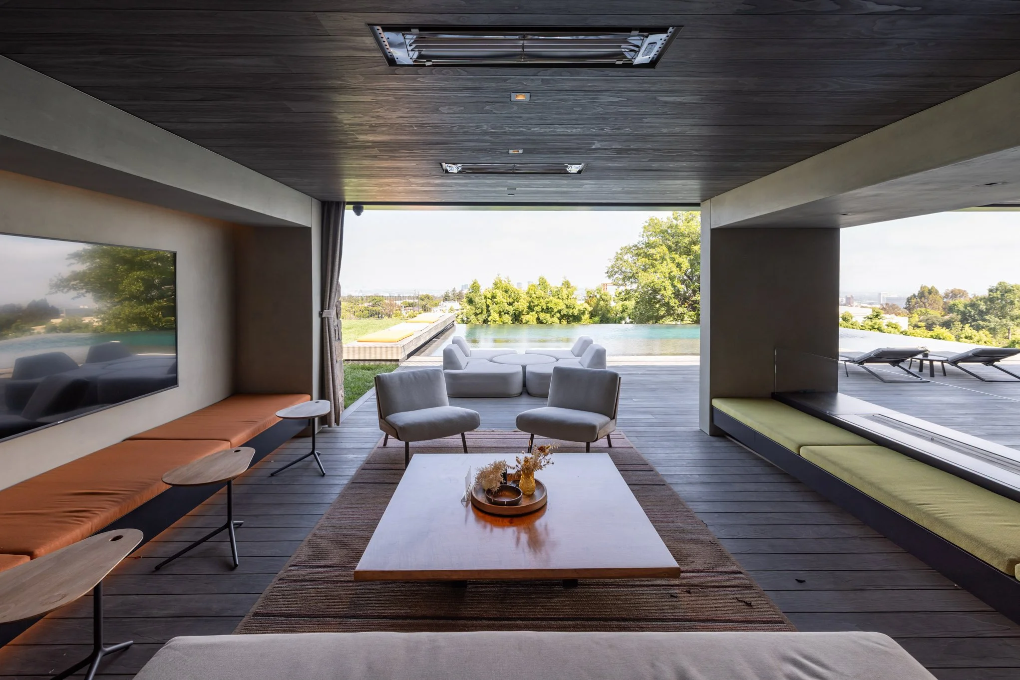 Living room with a view of a swimming pool and outdoor greenery, featuring modern furniture including gray armchairs, a large flat-screen TV, a wooden coffee table decorated with flowers, and a lounge area on the deck.