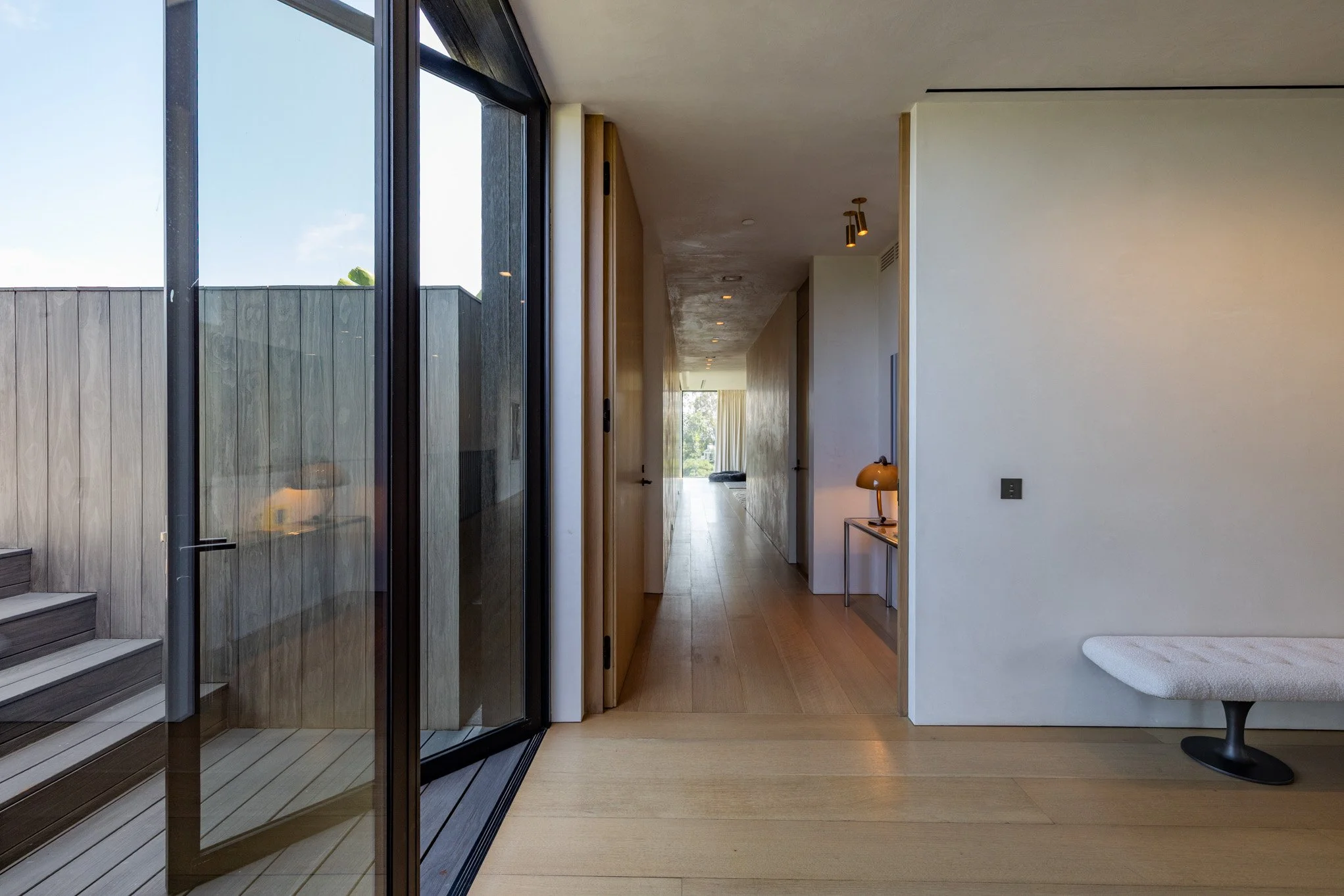 Interior view of a modern, minimalist home with wooden floors, a glass sliding door leading to an outdoor stairs, and a hallway with simple decor, including a small table with a lamp and a bench.