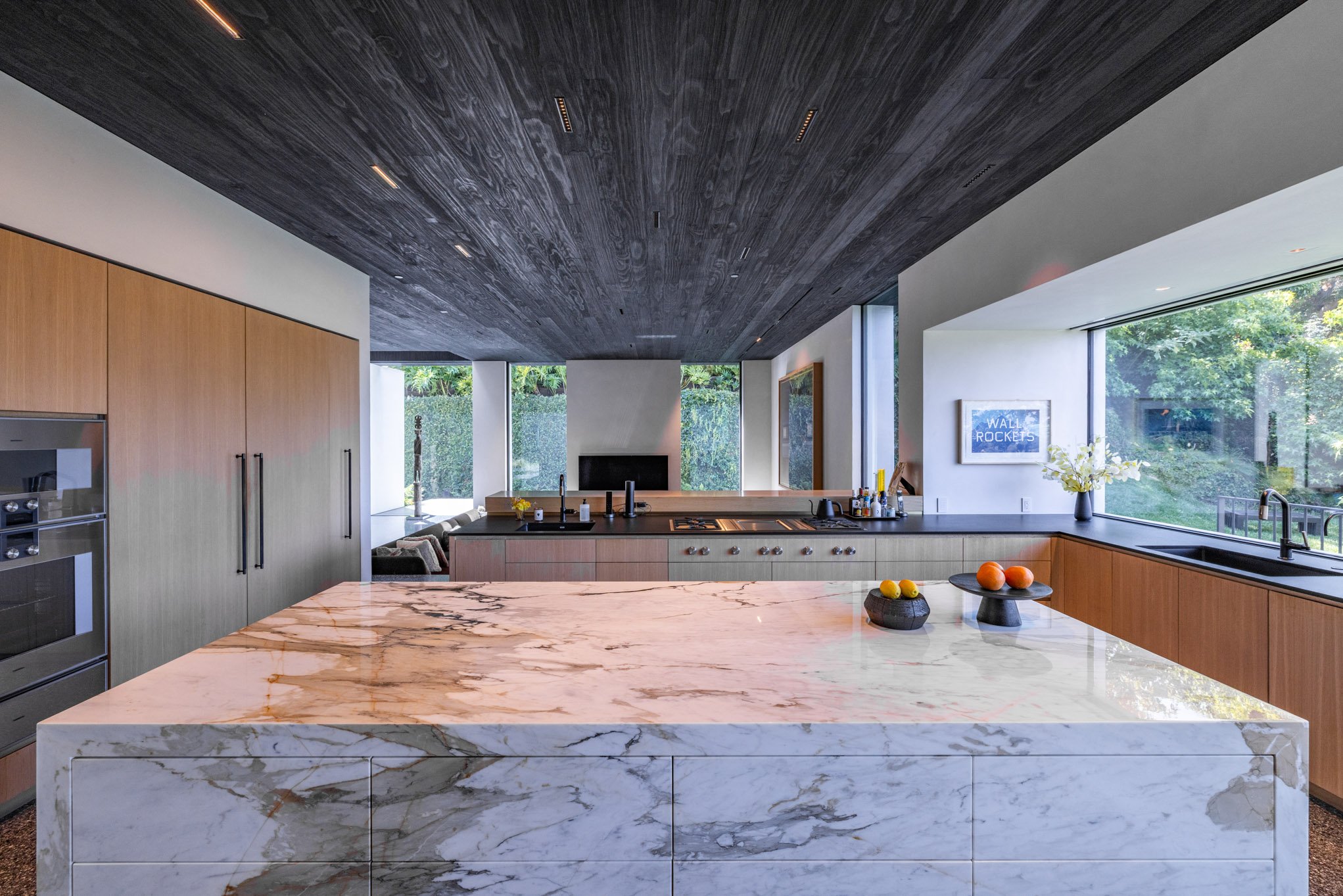 Modern kitchen with a marble island in the foreground, wooden cabinets on the left, large windows on the right, and a dark wood ceiling.