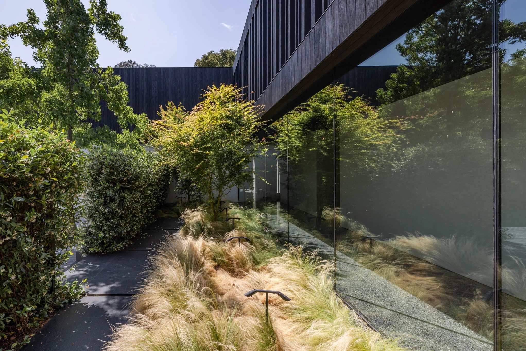 Modern building exterior with large glass windows, surrounded by greenery including trees, bushes, and ornamental grasses.