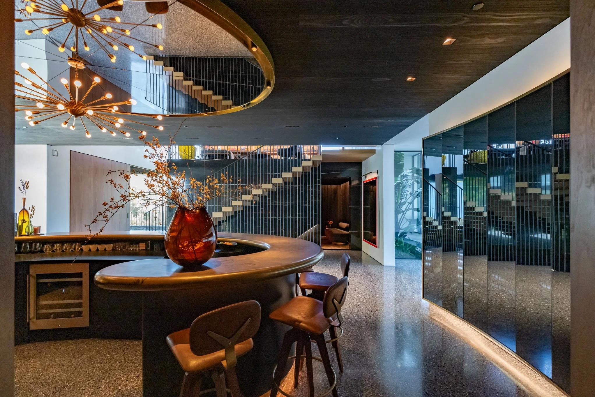 Modern hotel lobby with a curved bar, wooden chairs, a large floral arrangement in a red vase, reflective black wall panels, and a staircase with black railing leading upstairs.