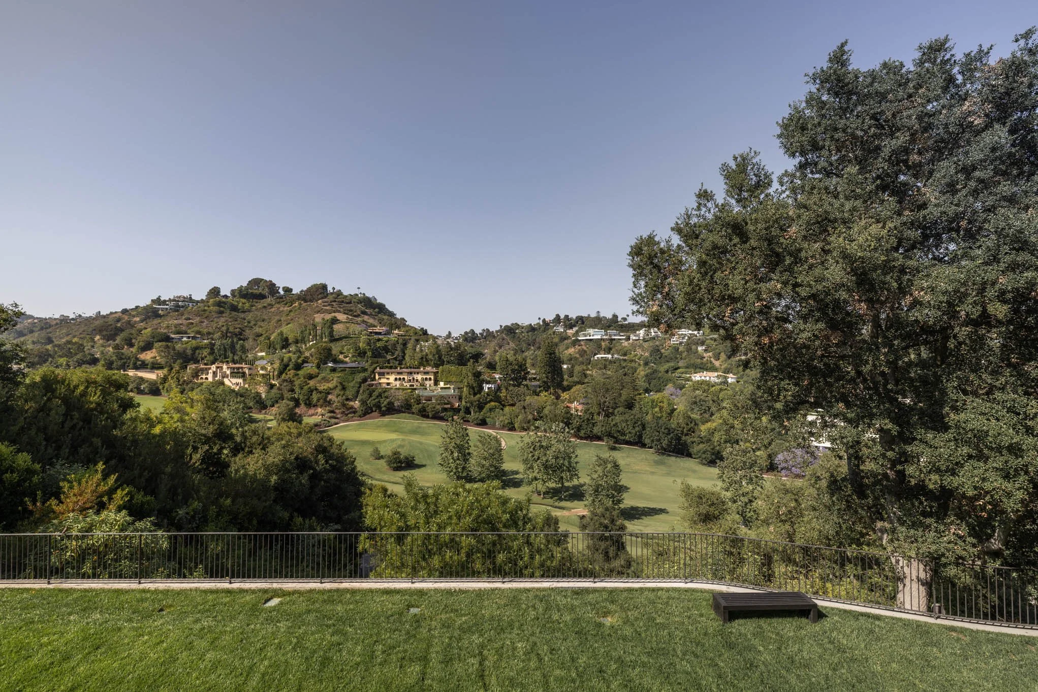 View of a hilly landscape with trees and buildings in the distance, a grassy area in the foreground, and a metal railing along the edge.