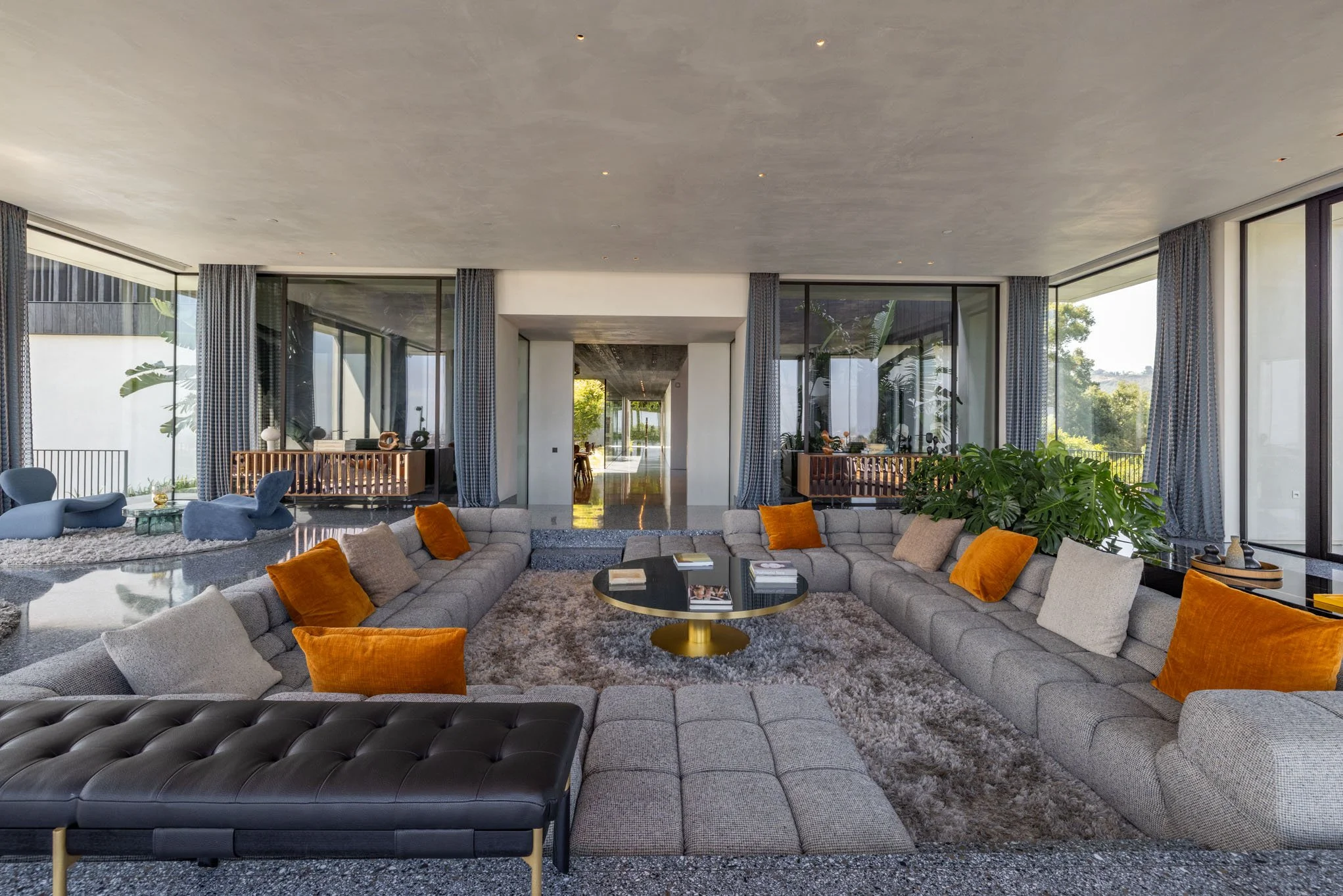 Modern living room with large gray sectional sofa, orange and beige pillows, a round black and gold coffee table, plush gray rug, large windows with curtains, and outdoor greenery visible through the windows.
