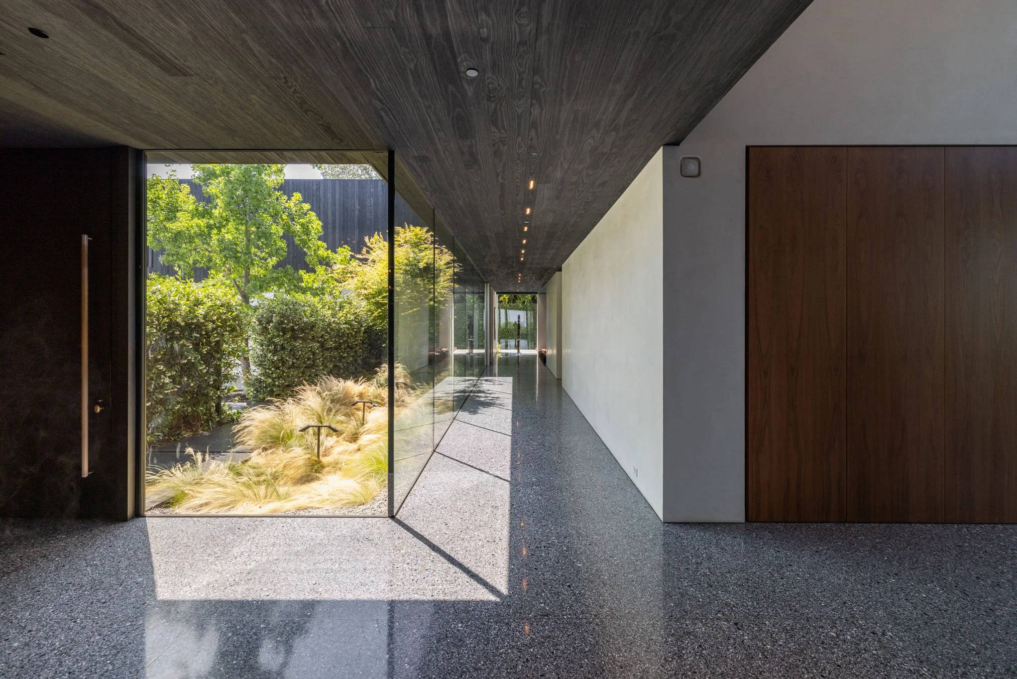 Modern indoor hallway with glass wall opening to a garden with bushes and trees, polished epoxy floor, dark wood ceiling, white wall on the right, and wooden panel on the far right.