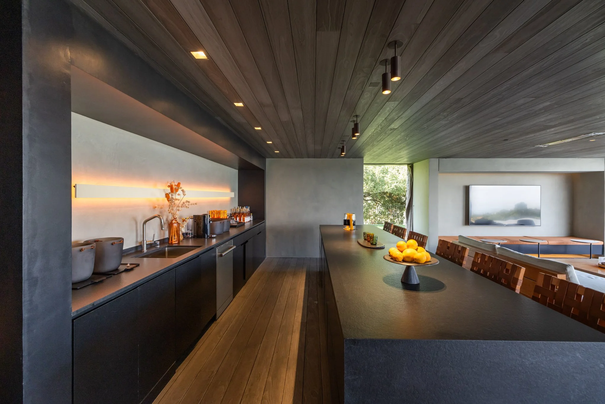 Modern kitchen area with a black countertop bar, serving station with glassware and fruit display, and a living room with a wall-mounted TV.