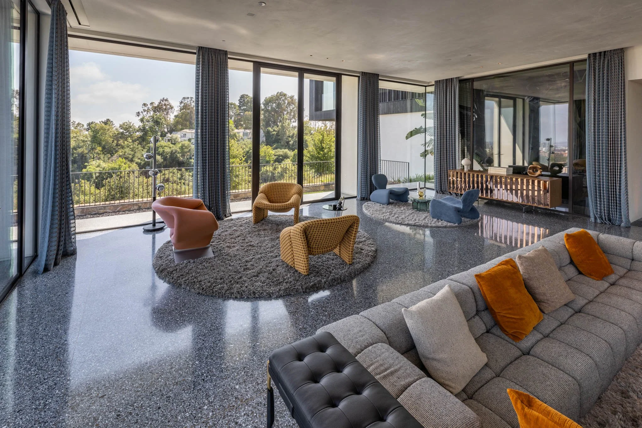 Modern living room with large glass windows overlooking greenery, featuring a gray sectional sofa with orange and beige pillows, four colorful armchairs on circular rugs, and a sleek wooden console table.