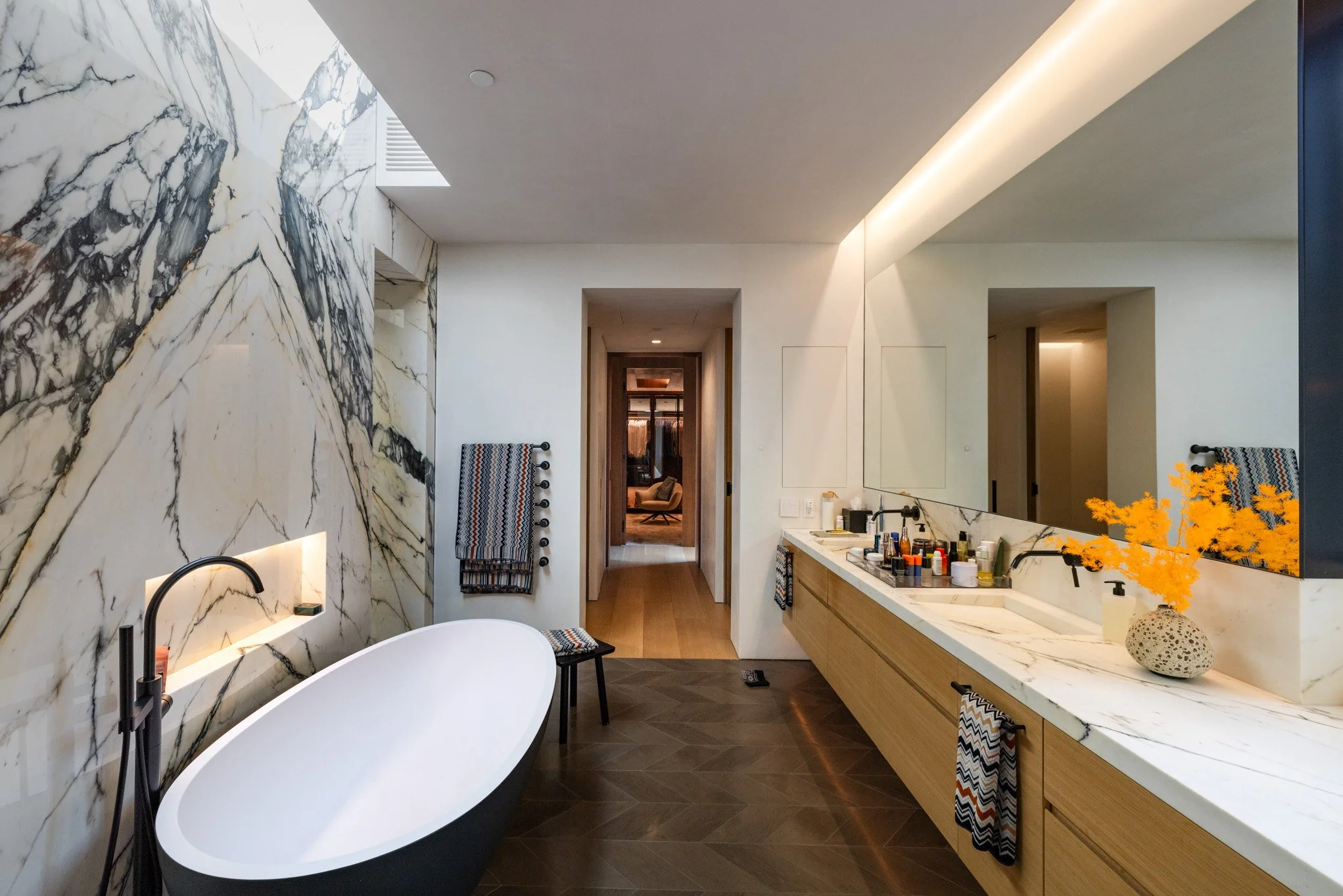 Modern bathroom with a freestanding bathtub, marble wall, wooden vanity with multiple toiletries, large mirror, and decorative orange flowers in a vessel. Towel hooks with patterned towels, wood flooring, and a distant view into a sitting area.