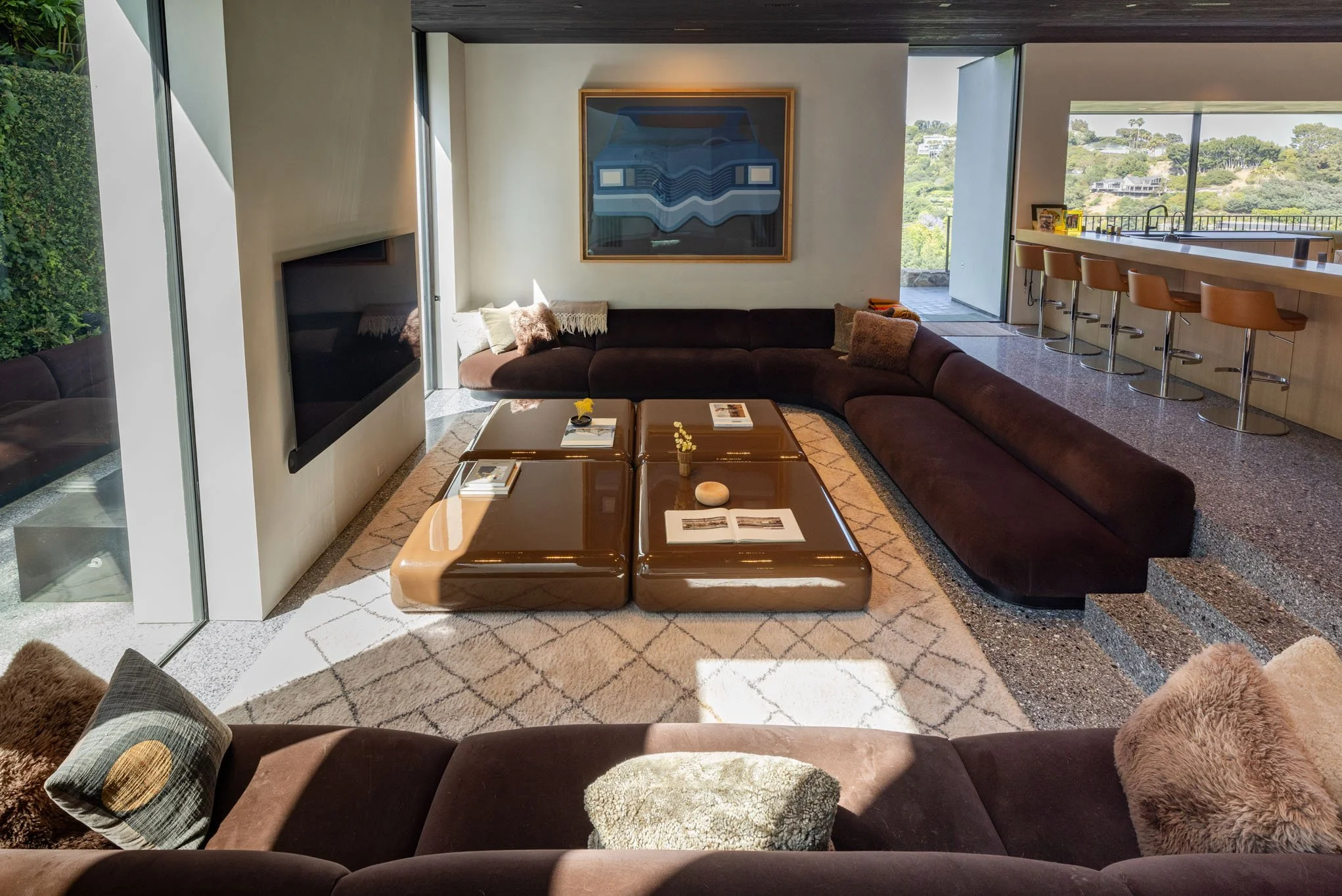 Living room with a large U-shaped dark brown sectional sofa, a modern glossy brown coffee table with books and decor, a wall-mounted TV, a large abstract painting, a patterned area rug, and floor-to-ceiling windows showing an outdoor landscape with g