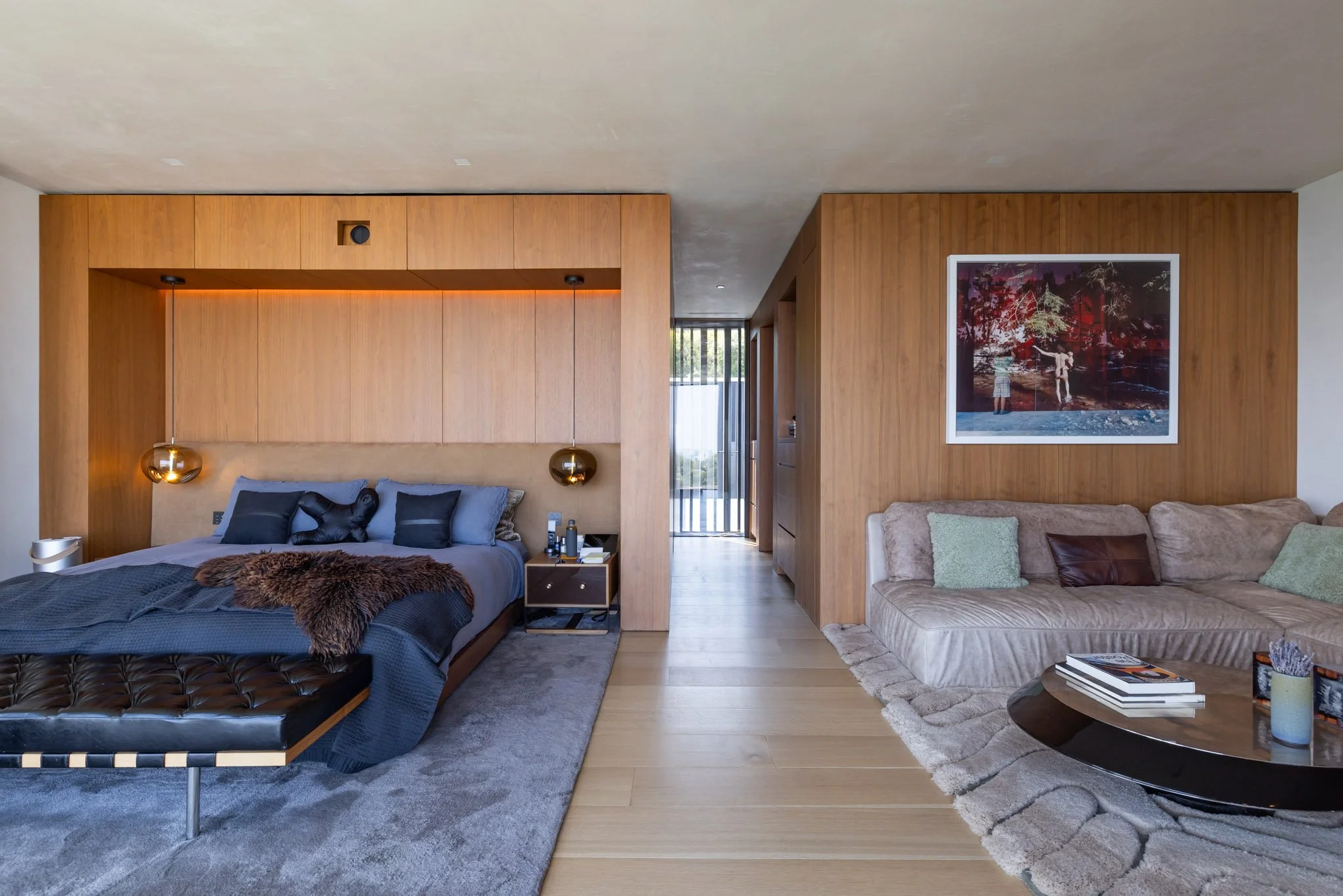 Hotel bedroom with a bed on the left, featuring black and gray bedding and a furry throw, and a beige sectional sofa on the right with pillows, a coffee table with books, and a framed picture on the wooden wall background.