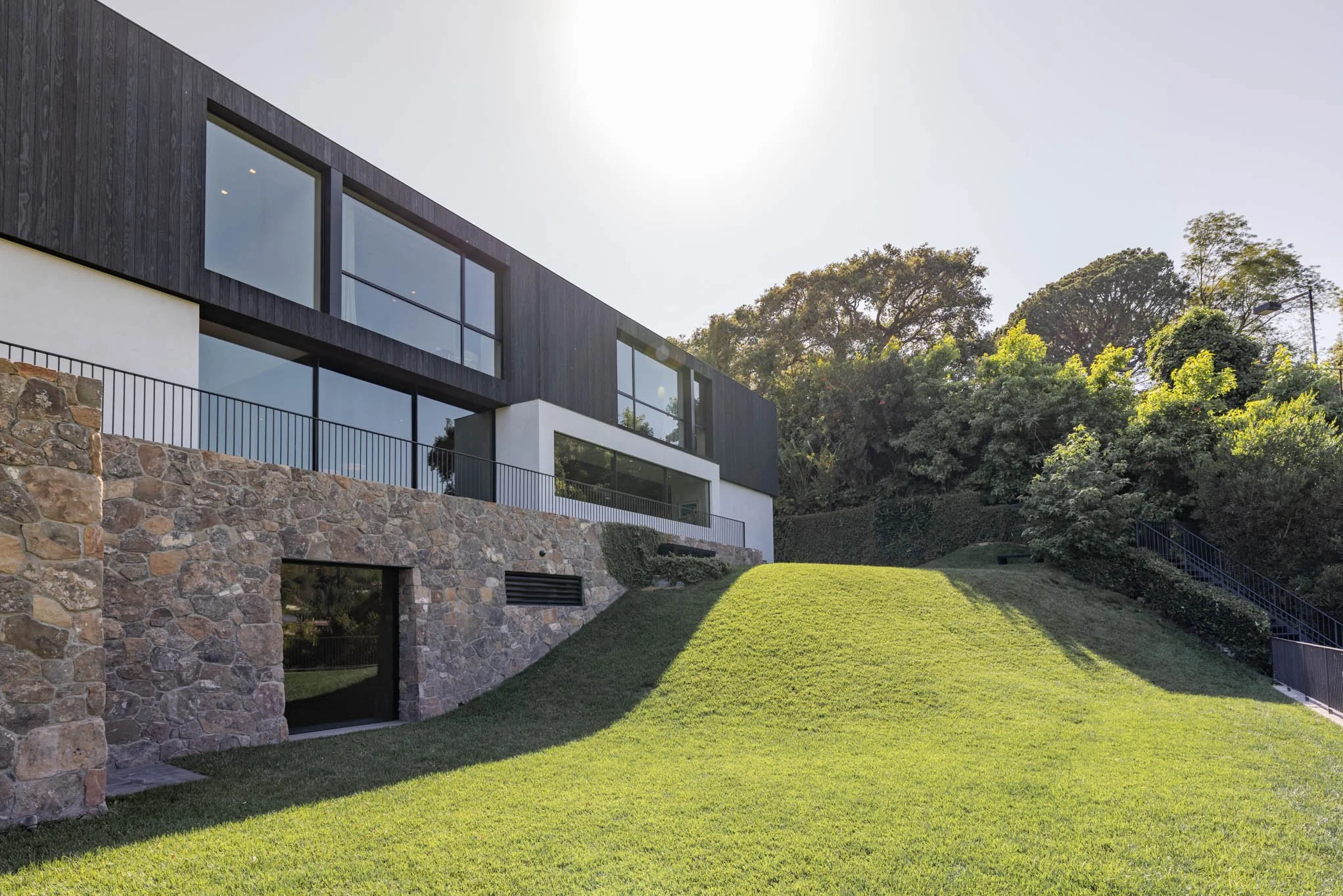 Modern house with large windows, stone foundation, and black cladding, situated on a grassy hill with trees in the background under a clear sky.