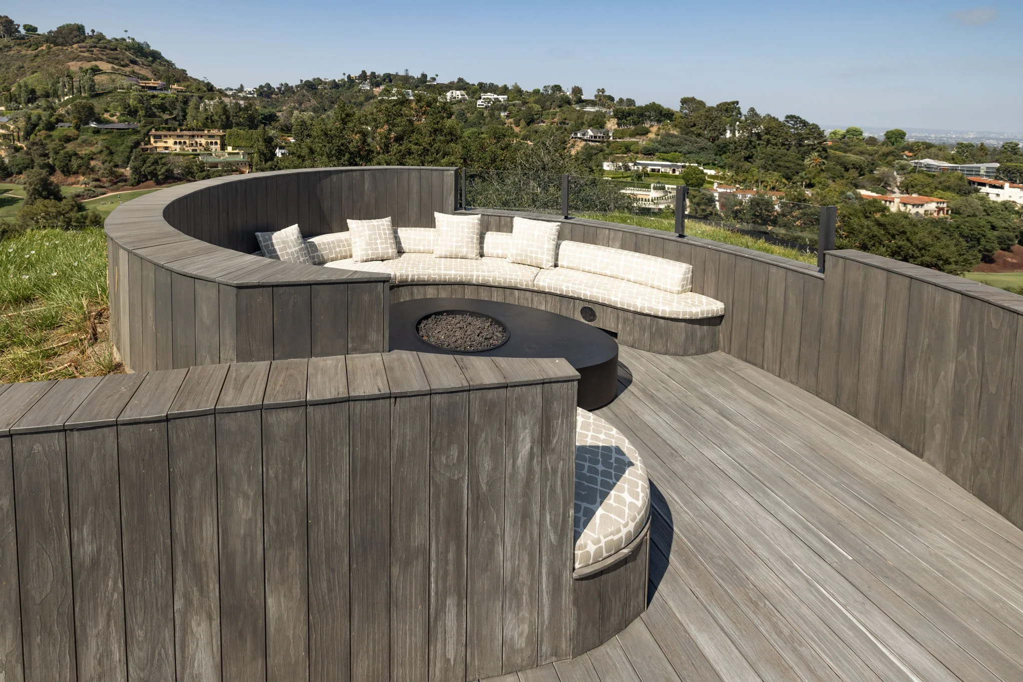 Outdoor modern curved wooden decking with built-in seating area and patterned cushions, overlooking a hilly landscape with homes and greenery.