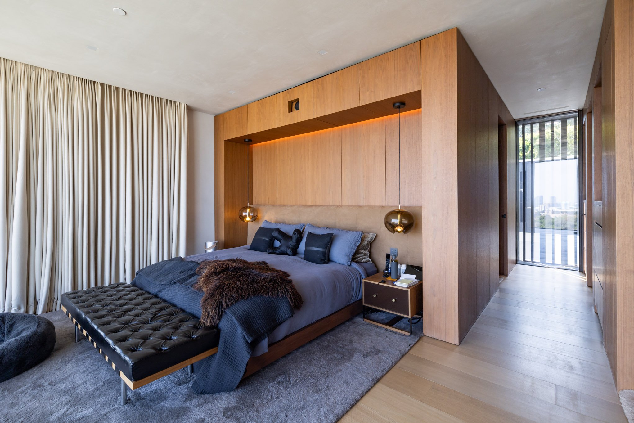 Modern bedroom with a wooden headboard, black and gray bedding, pendant lights, a small nightstand with books, and large cream curtains.