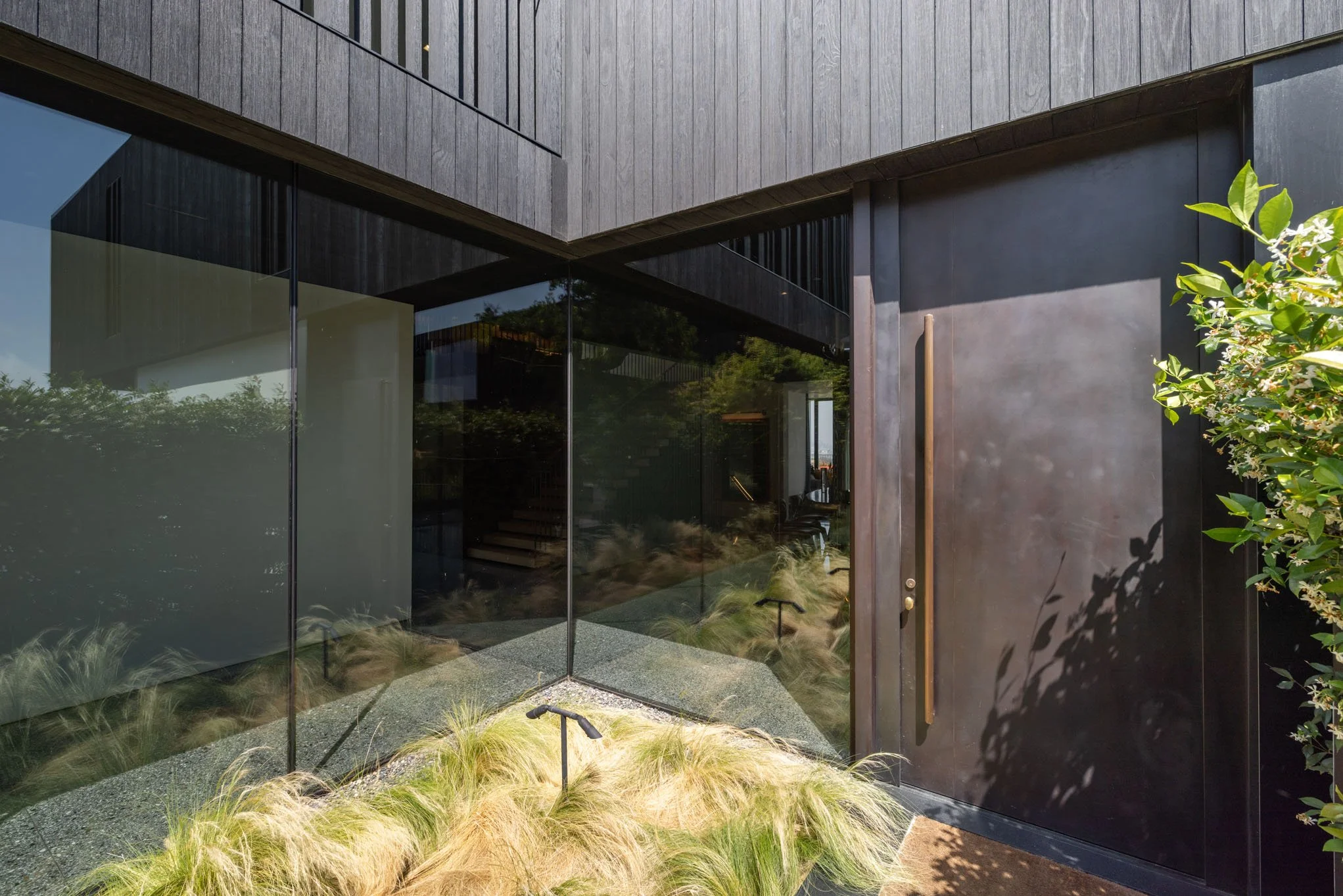 Modern building entrance with large glass windows, dark metal door, and landscaped greenery.