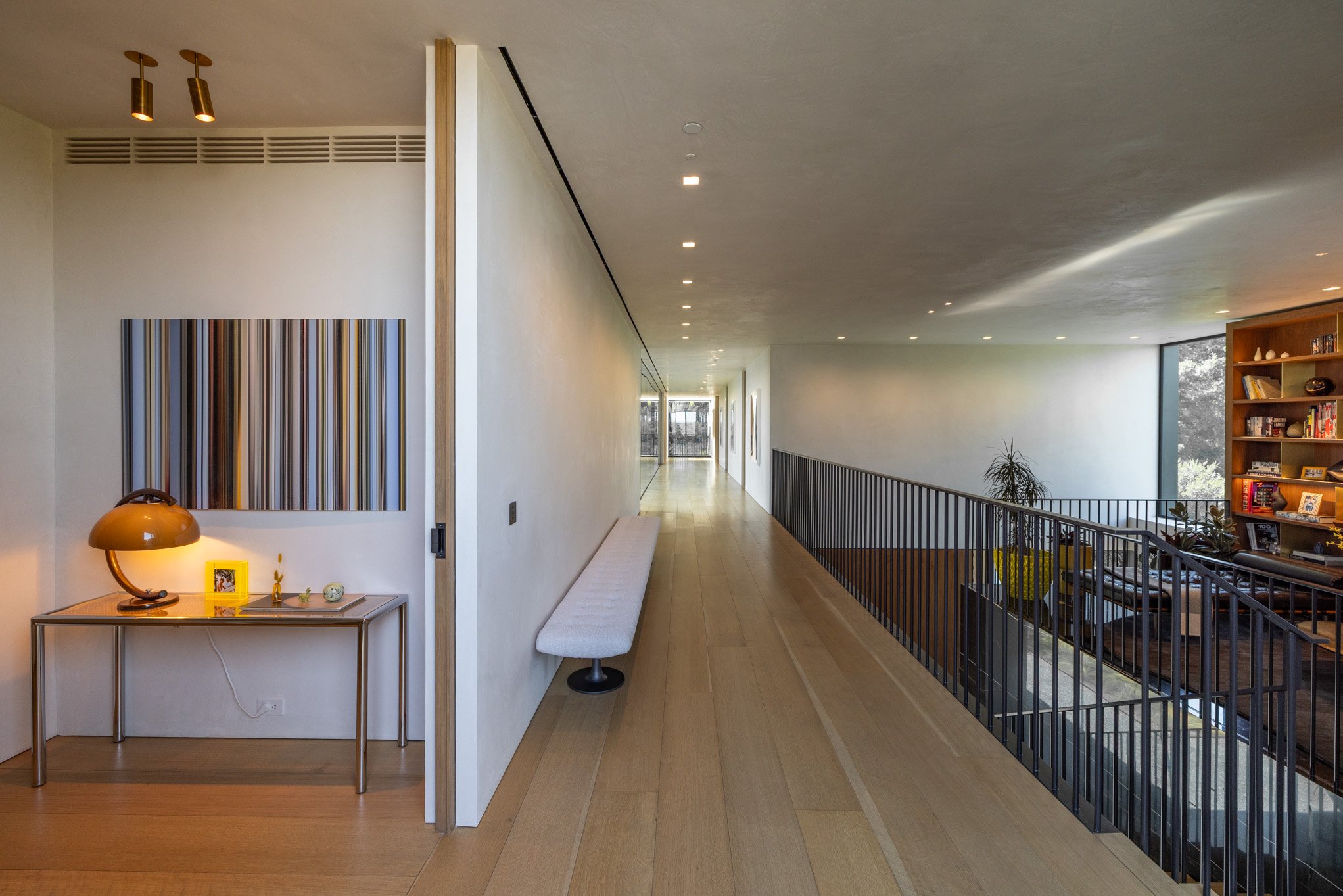 Modern interior hallway with wooden flooring, a long white bench on the left, artwork on the wall, and a staircase with black railing leading to a living space with a bookshelf and large windows showing trees outside.