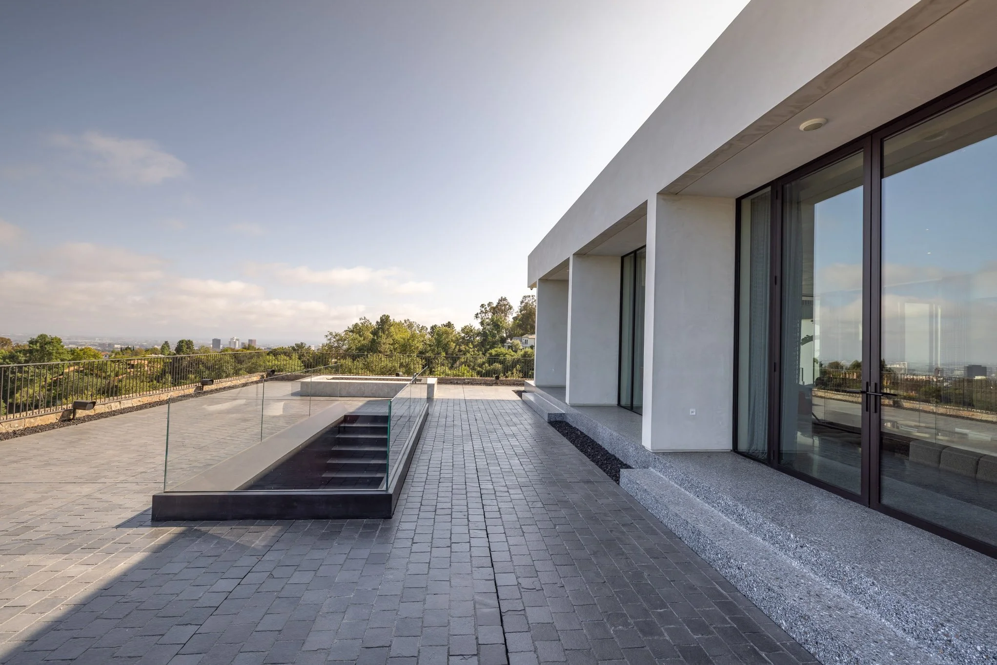 Modern building patio with gray pavers, glass railing, and sliding glass doors, overlooking a landscape with trees and a partly cloudy sky.
