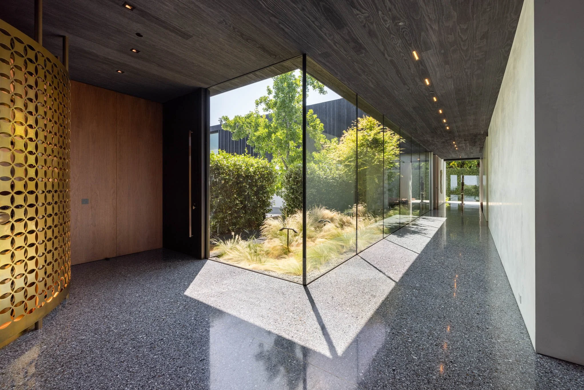 Modern interior hallway with large glass windows revealing a lush green garden outside, polished concrete floors, a dark wood ceiling with recessed lighting, and a decorative gold partition on the left side.