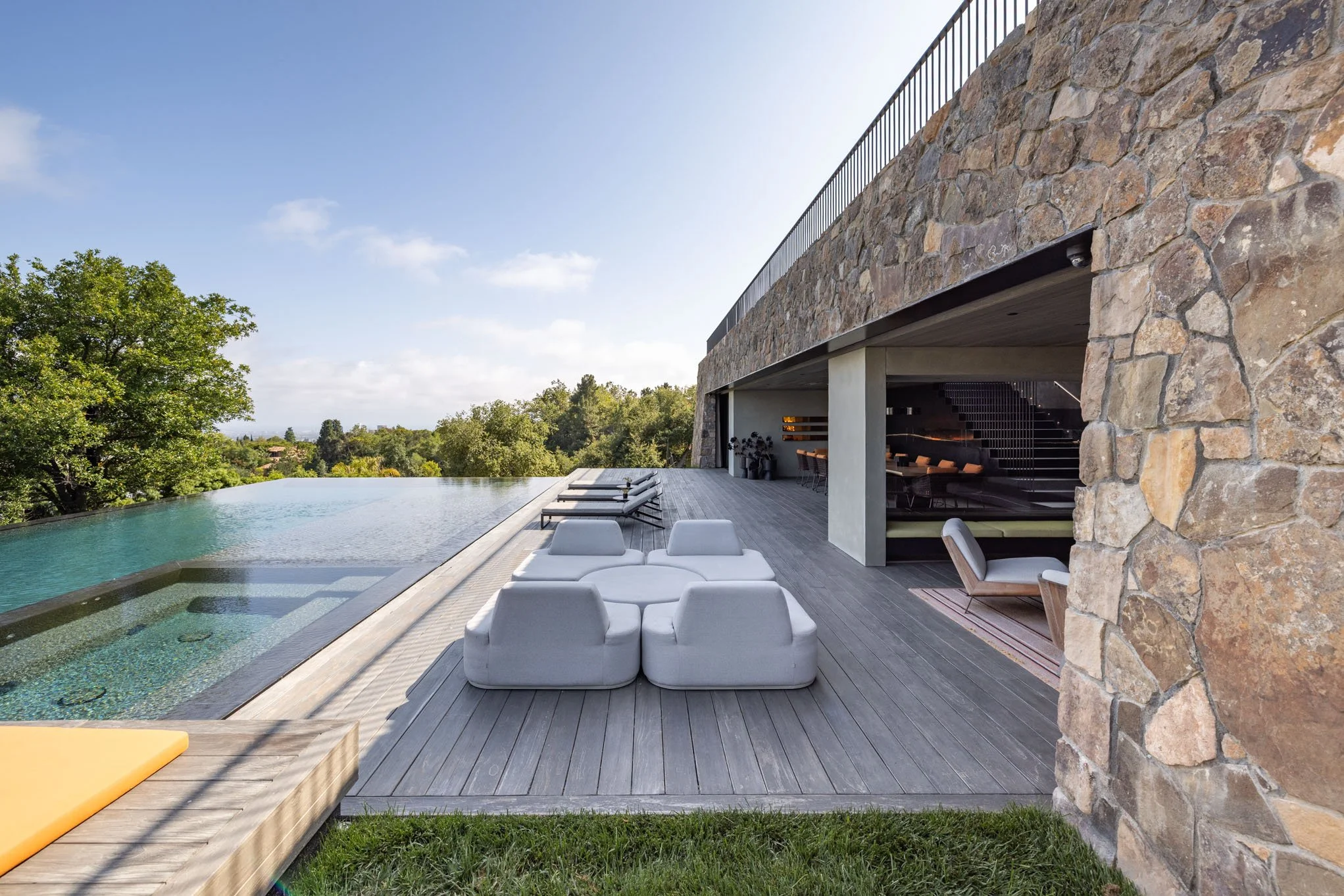 Modern house with outdoor living area, pool, and infinity edge on a sunny day, surrounded by trees.