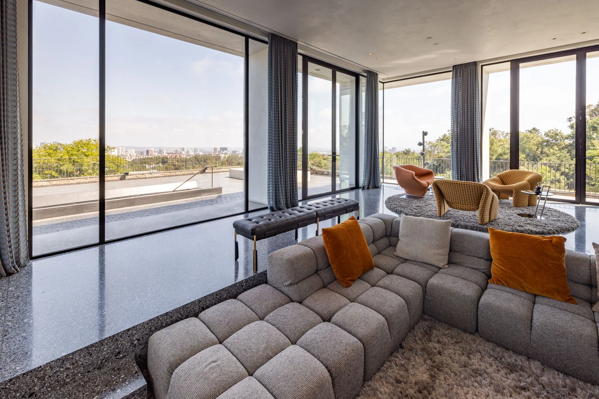 Modern living room with large floor-to-ceiling windows, view of city skyline, gray sectional sofa with orange pillows, upholstered chairs, rug, and balcony.